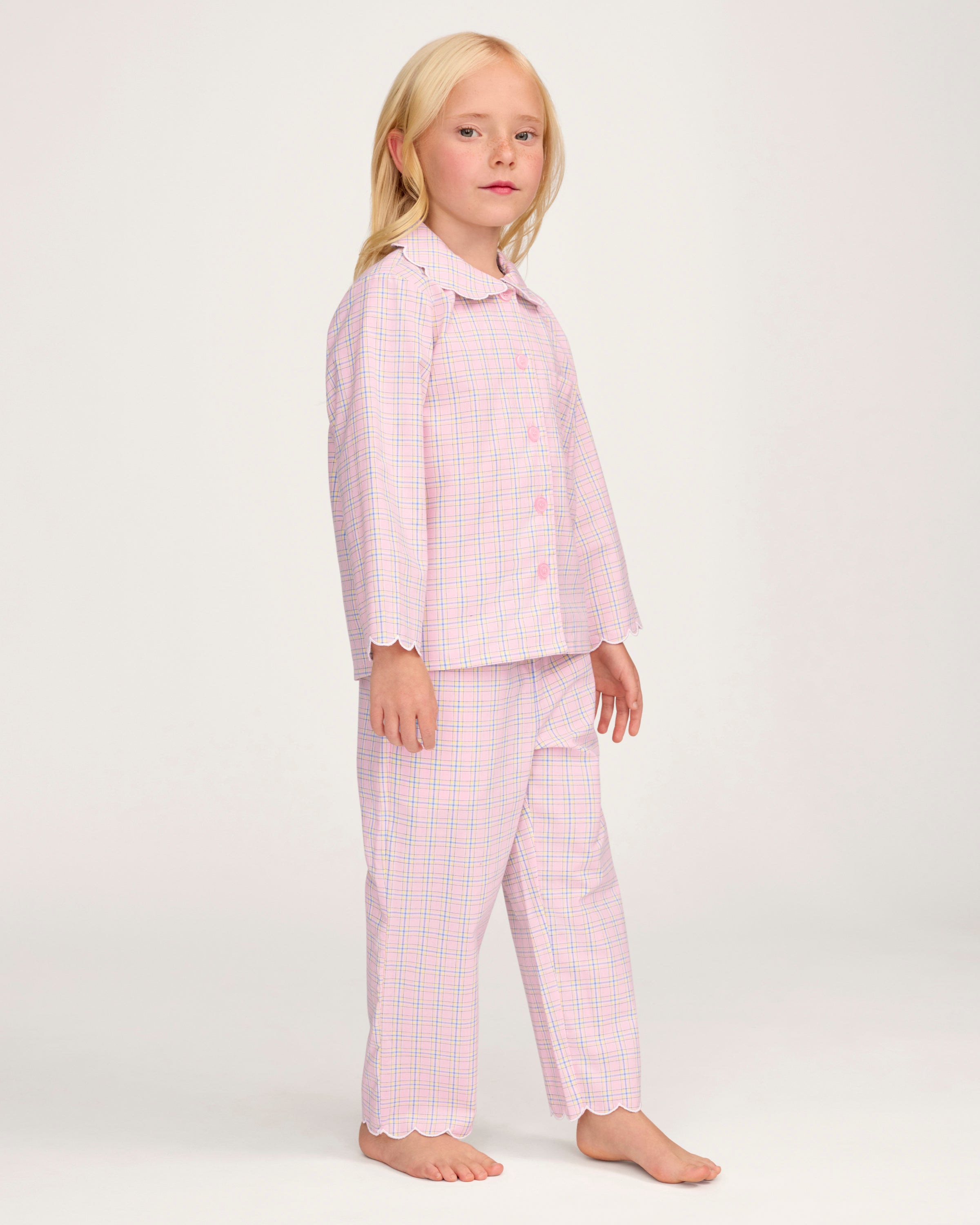 Young girl with long blonde hair wears the Kid's Twill Colette Pajama Set in Peony Plaid, featuring pink and white checks, scalloped edges, and stands barefoot against a plain light background.
