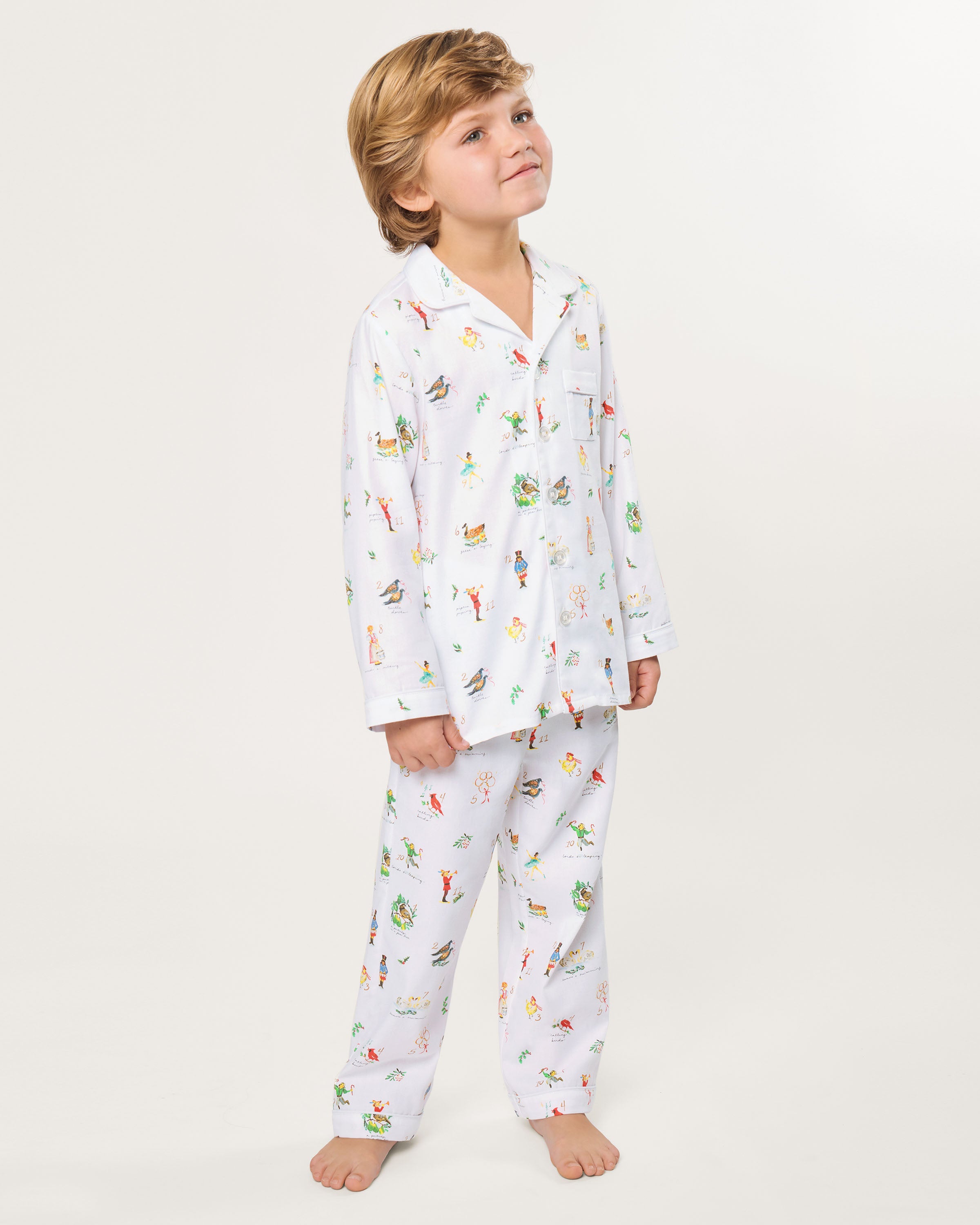 The Kid's Twill Pajama Set in 12 Days of Christmas features a white, long-sleeve button-up top with collar and front pocket, plus matching pants, all adorned with playful, colorful holiday illustrations of people and animals.