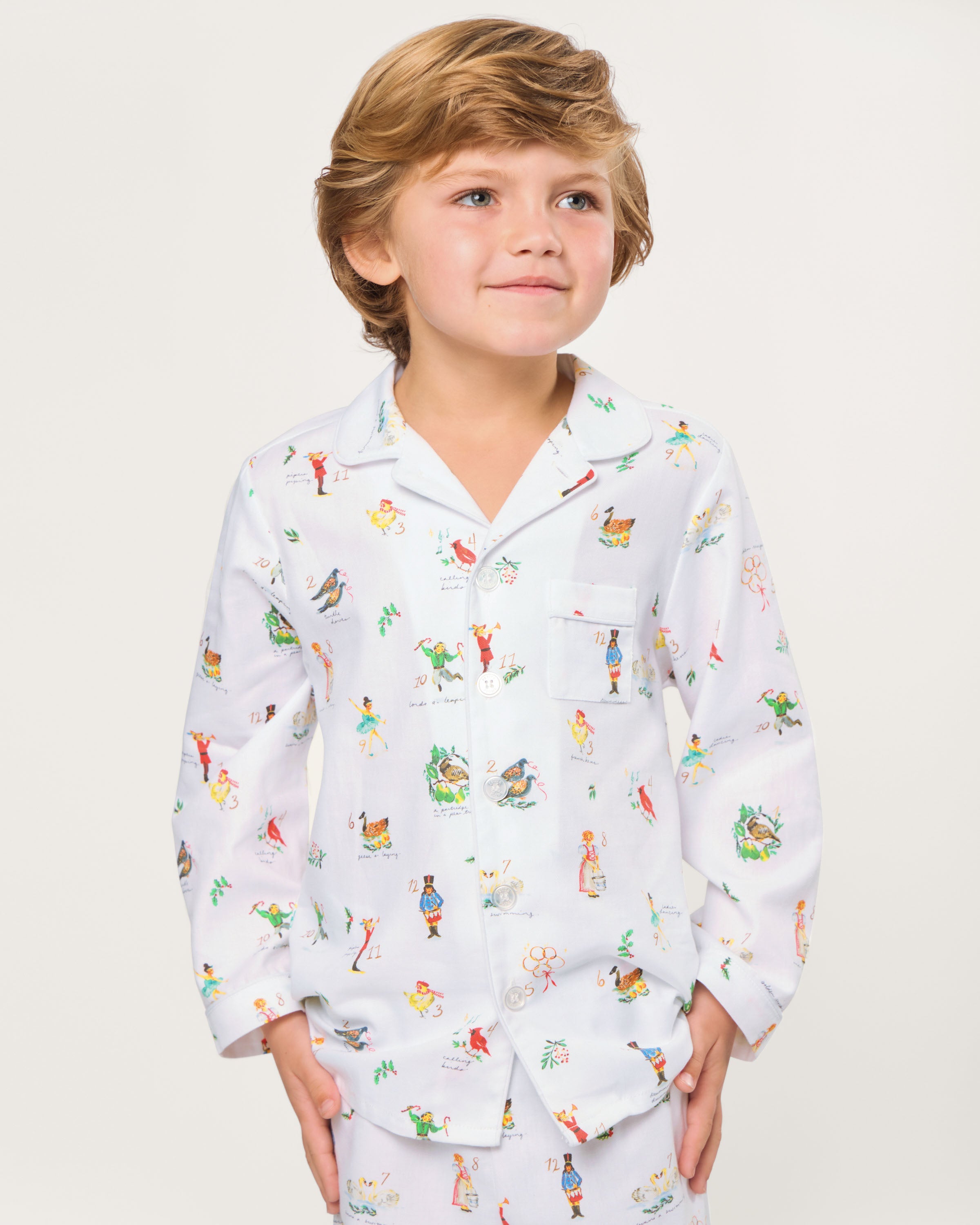 The Kid's Twill Pajama Set in 12 Days of Christmas features a white, long-sleeve button-up top with collar and front pocket, plus matching pants, all adorned with playful, colorful holiday illustrations of people and animals.