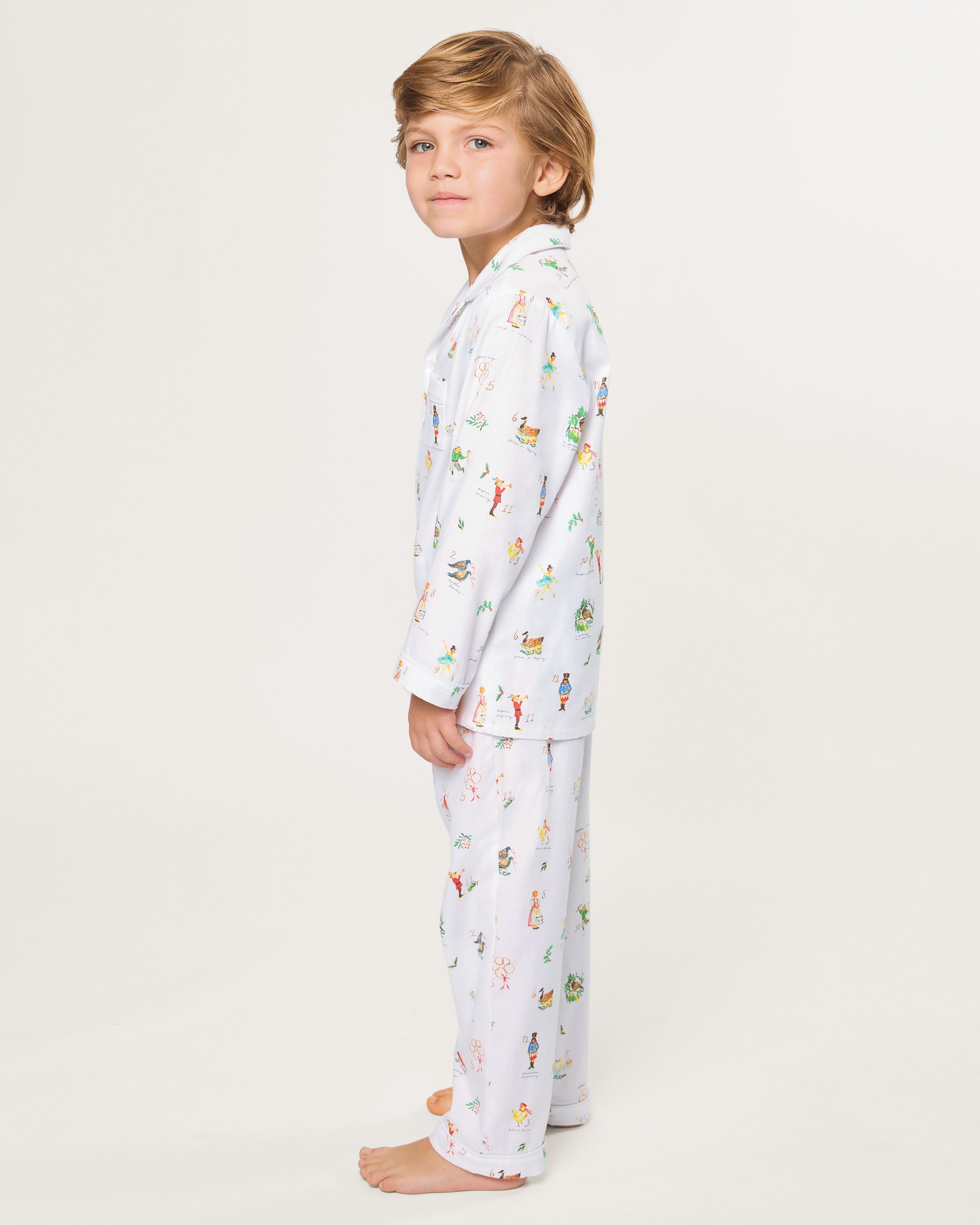 The Kid's Twill Pajama Set in 12 Days of Christmas features a white, long-sleeve button-up top with collar and front pocket, plus matching pants, all adorned with playful, colorful holiday illustrations of people and animals.