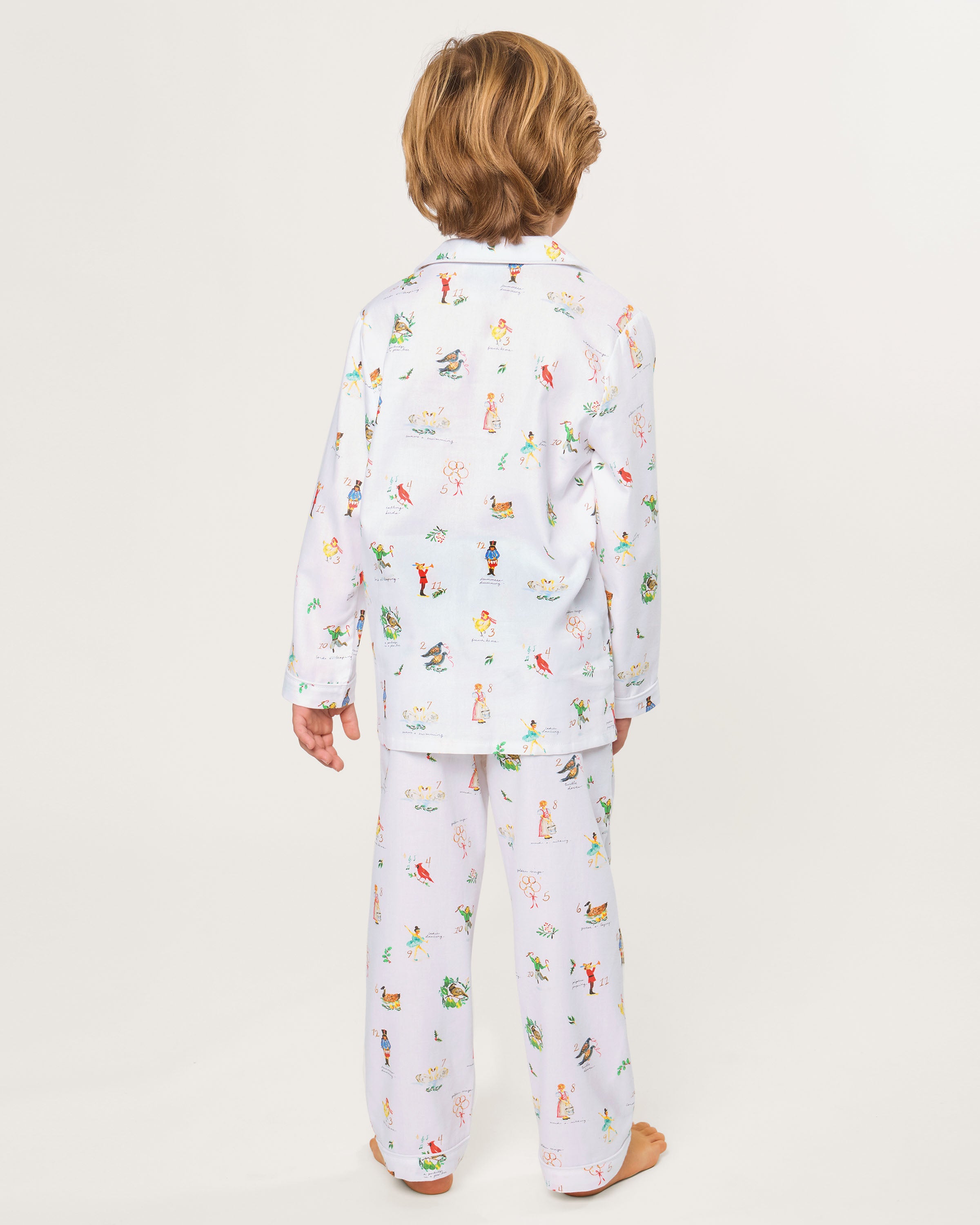 The Kid's Twill Pajama Set in 12 Days of Christmas features a white, long-sleeve button-up top with collar and front pocket, plus matching pants, all adorned with playful, colorful holiday illustrations of people and animals.
