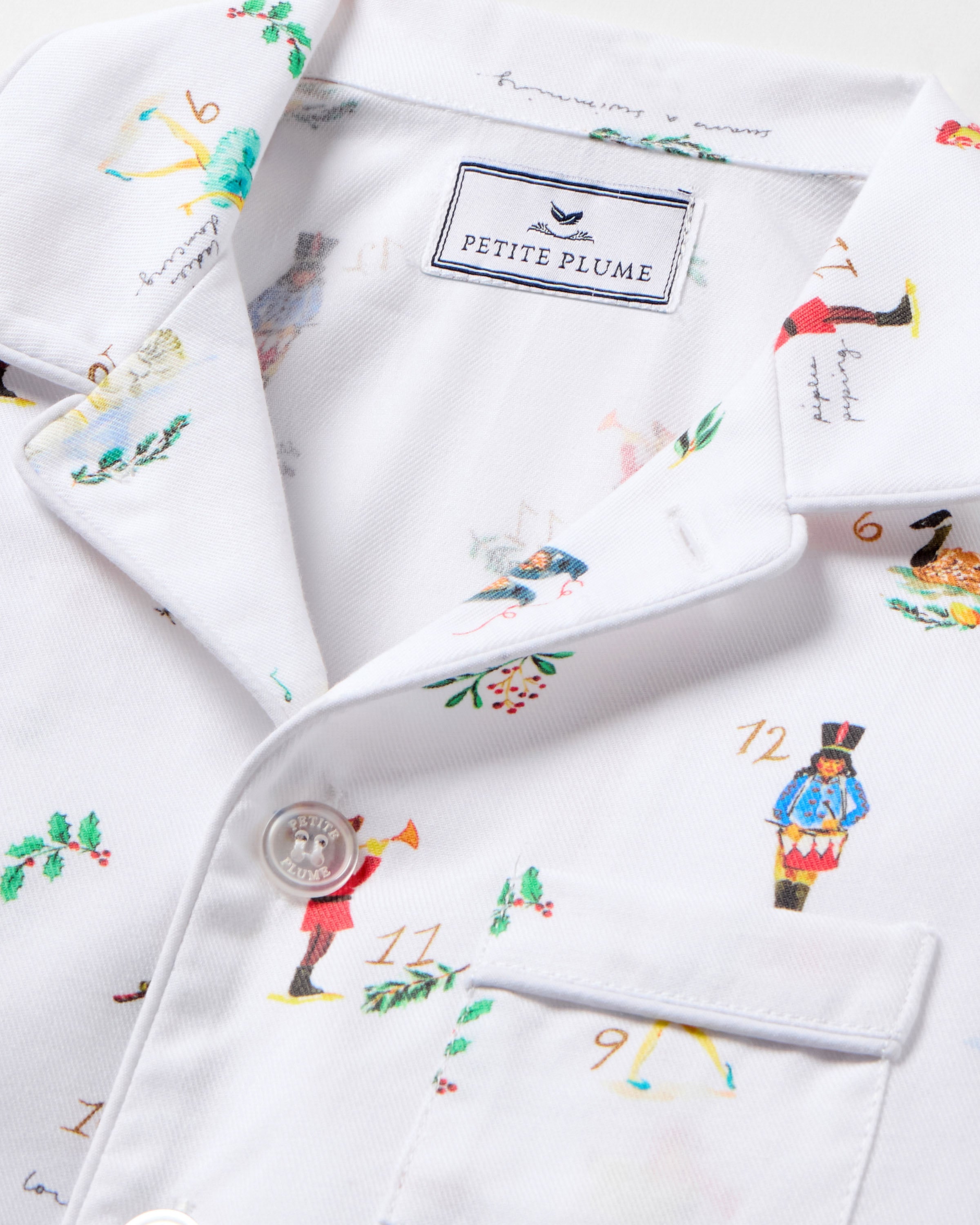 The Kid's Twill Pajama Set in 12 Days of Christmas features a white, long-sleeve button-up top with collar and front pocket, plus matching pants, all adorned with playful, colorful holiday illustrations of people and animals.