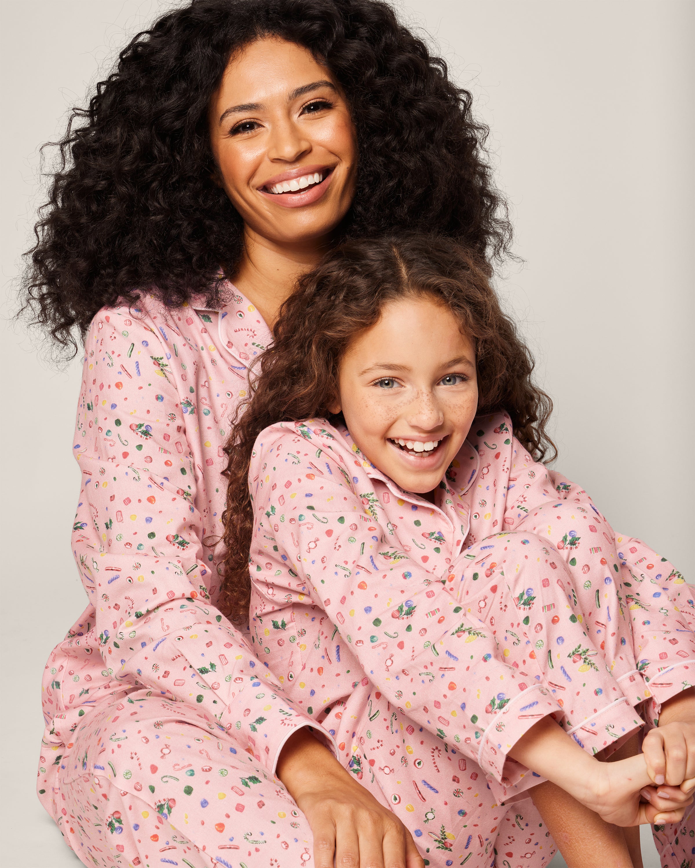 A smiling woman and young girl sit together, both in matching Petite Plumes Womens Twill Pajama Set in Vintage Sweets. The woman lovingly embraces the girl as they happily pose against a plain background, their outfits reminiscent of delightful holiday treats.