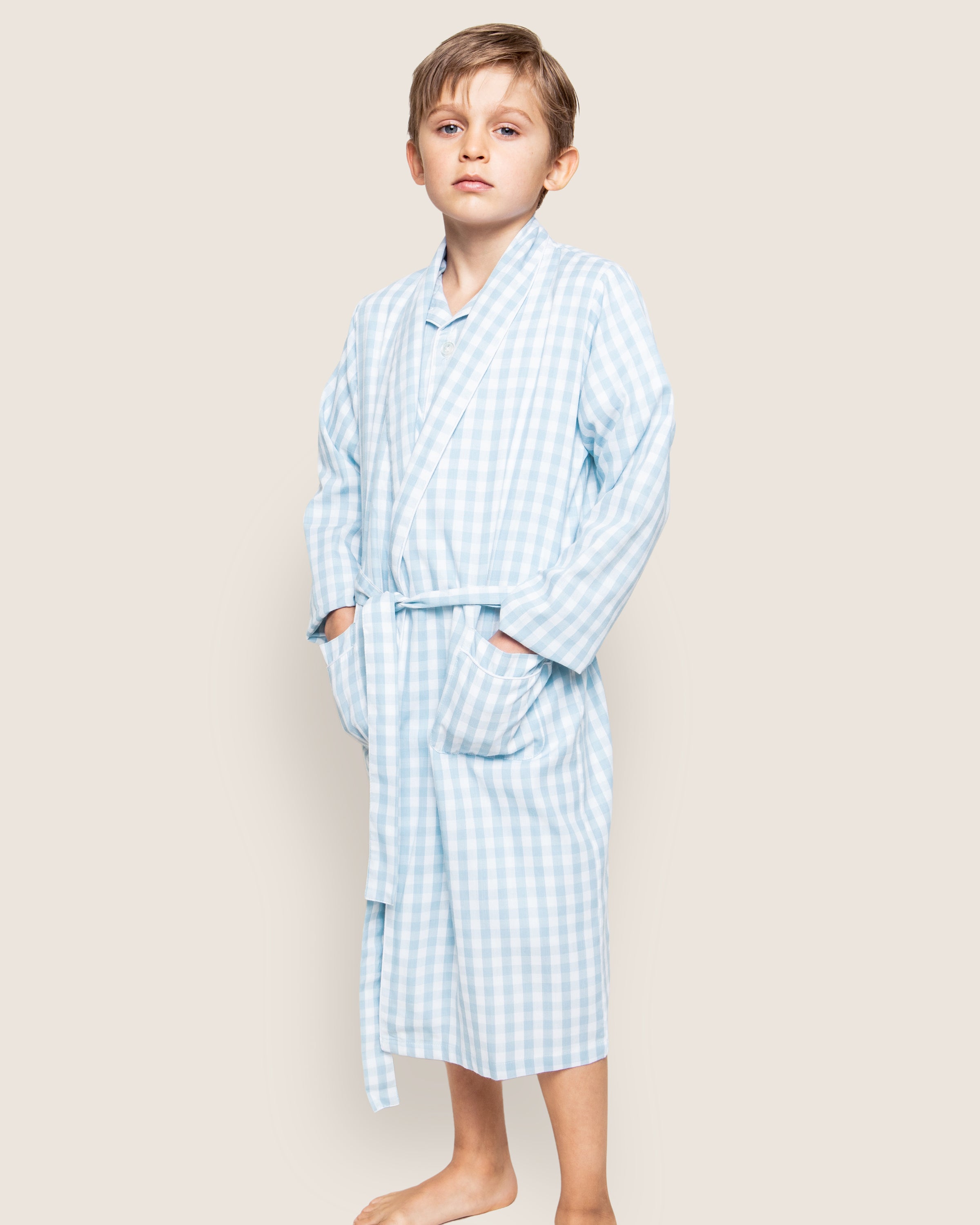 Against a beige background, a young boy stands barefoot in Petite Plumes Kids Twill Robe in Light Blue Gingham. Made from flame-retardant fabric and featuring pockets, he embodies safe and stylish childrens sleepwear as he looks directly at the camera.