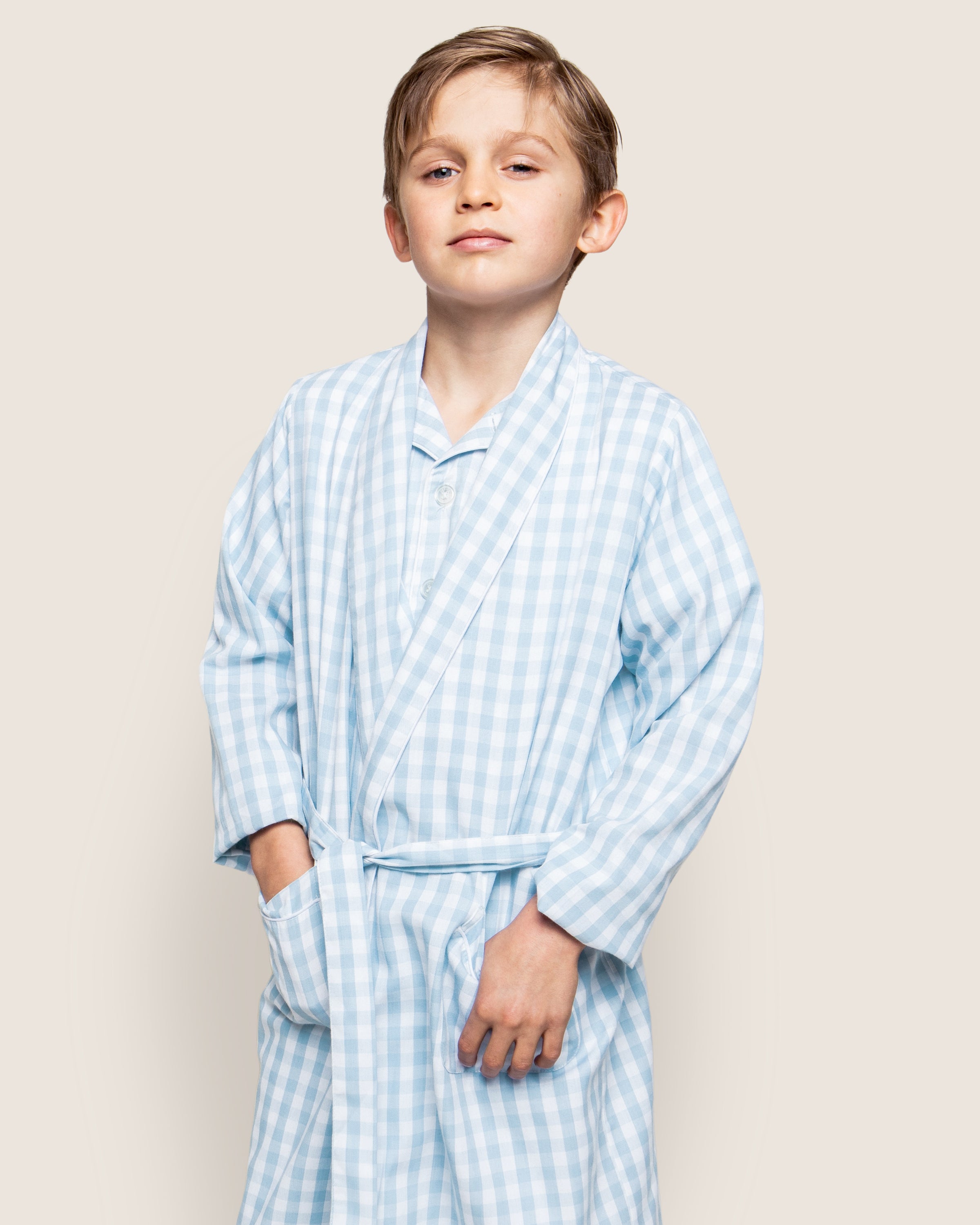 A young boy confidently poses in Petite Plumes Kids Twill Robe in Light Blue Gingham, with his hands in the pockets and a relaxed expression against a neutral light background.