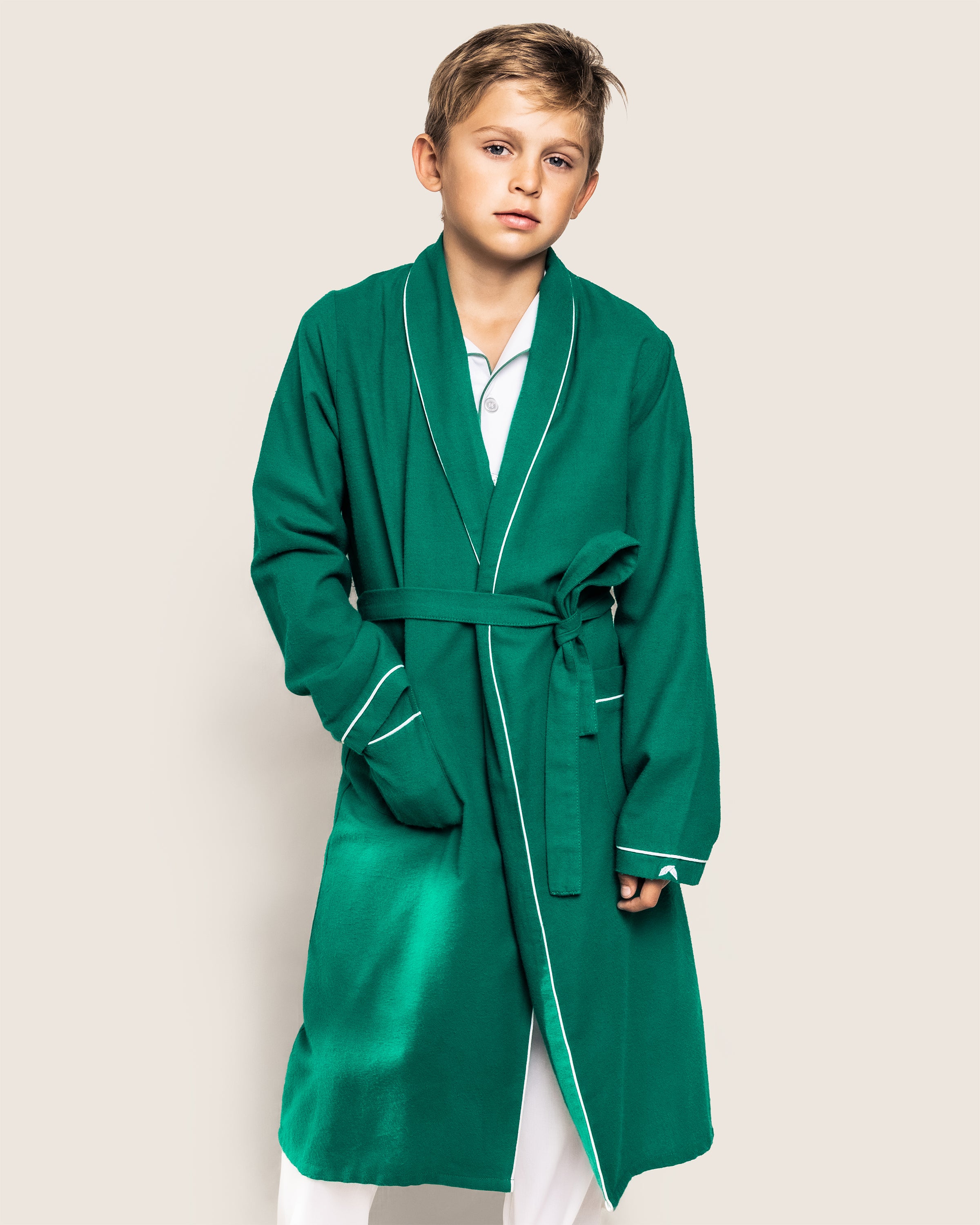 A young boy models the Petite Plume Kids Flannel Robe in Forest Green with White Piping against a plain background, looking at the camera with one hand in his pocket. The flame-retardant robe tied at the waist reveals white pants, showcasing cozy childrens sleepwear.