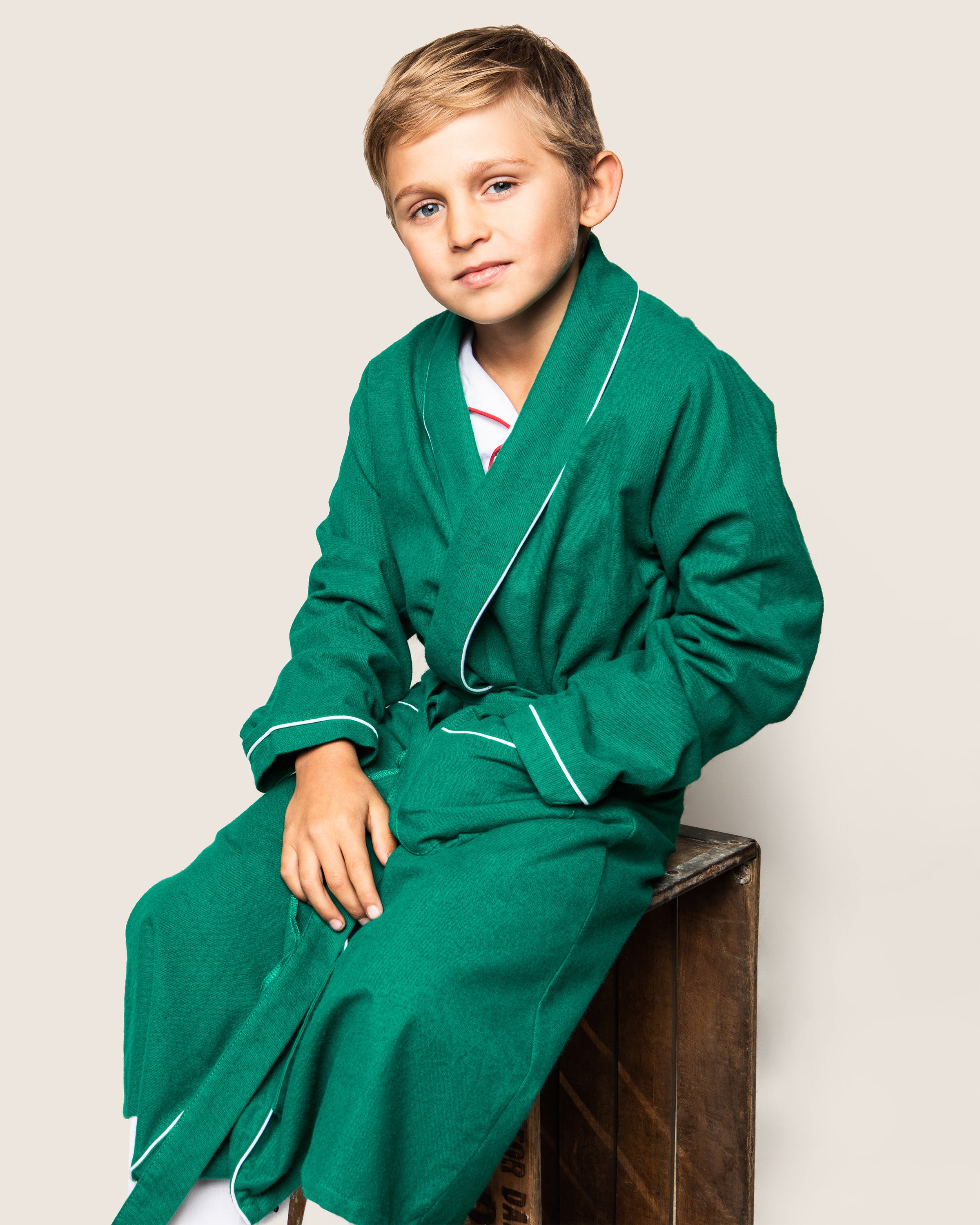 A blond boy sits on a wooden box in a Petite Plume Kids Flannel Robe, forest green with white piping, over a white outfit against an off-white background complementing his cozy ensemble.