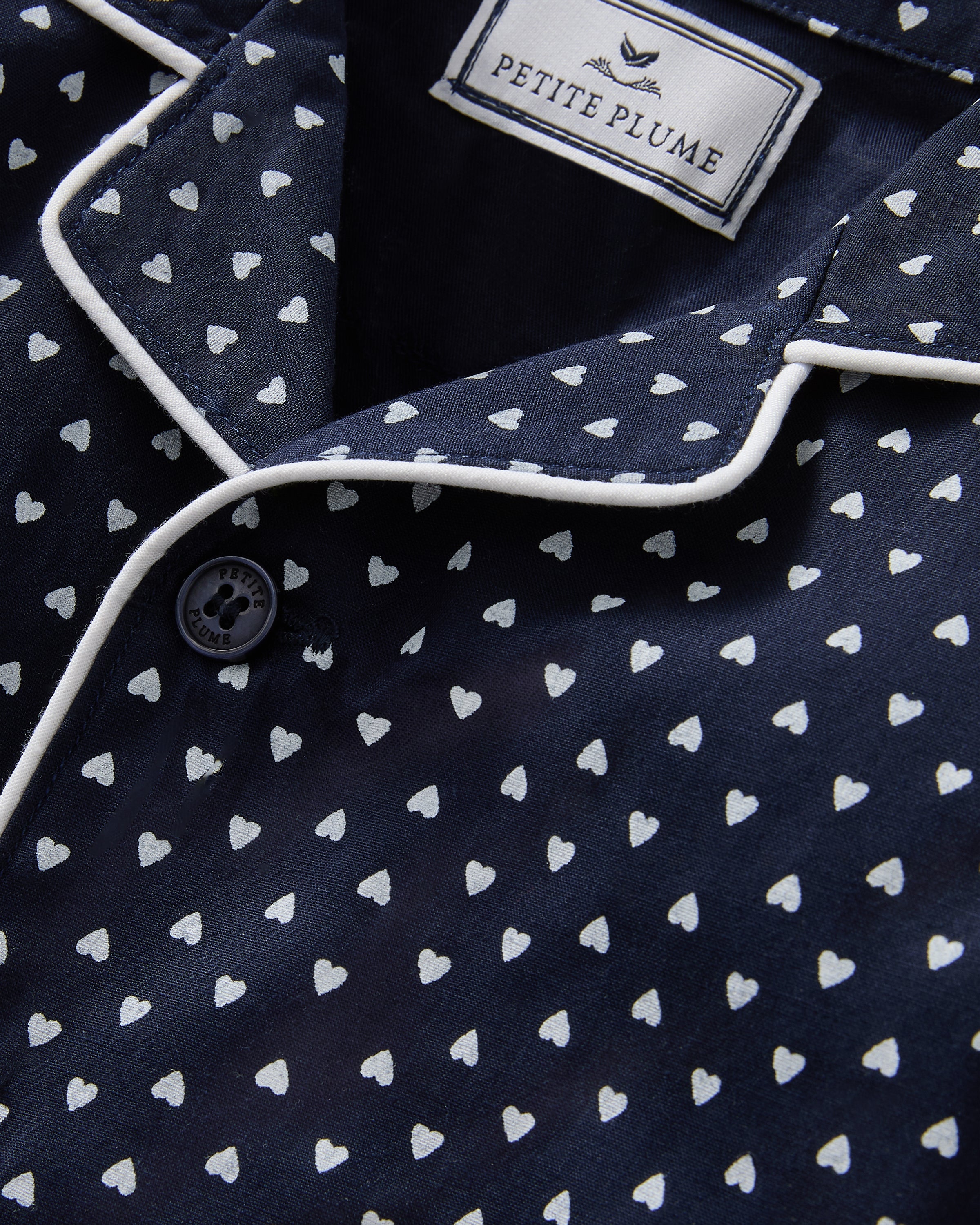 The Baby's Twill Classic Romper in Midnight Hearts is a navy blue onesie with long sleeves and legs, featuring a white heart pattern and white piping on the collar, cuffs, and front.