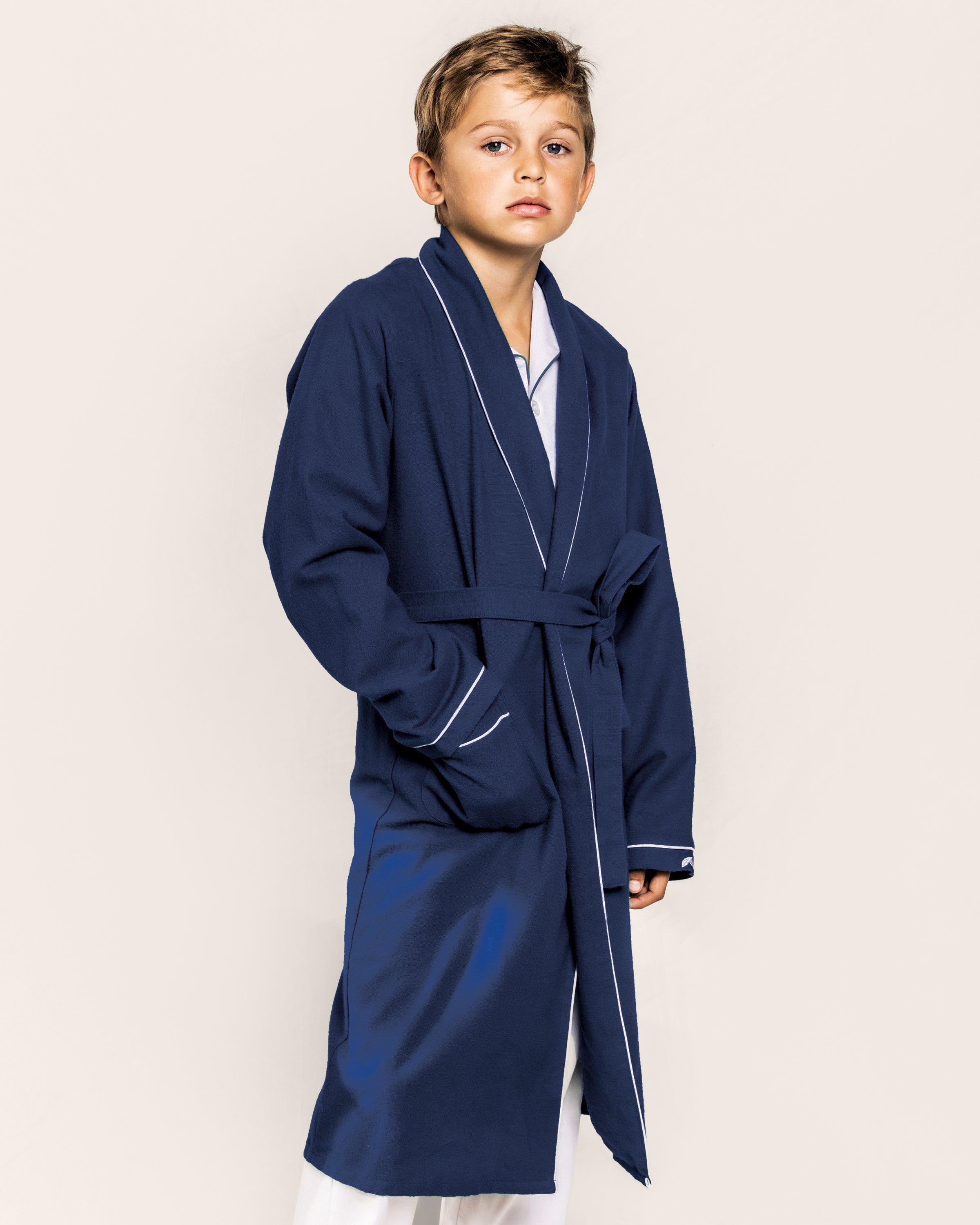 A young boy wears the Petite Plume Kids Flannel Robe in Navy with White Piping, belted over flame-retardant white pajamas, standing against a light background.