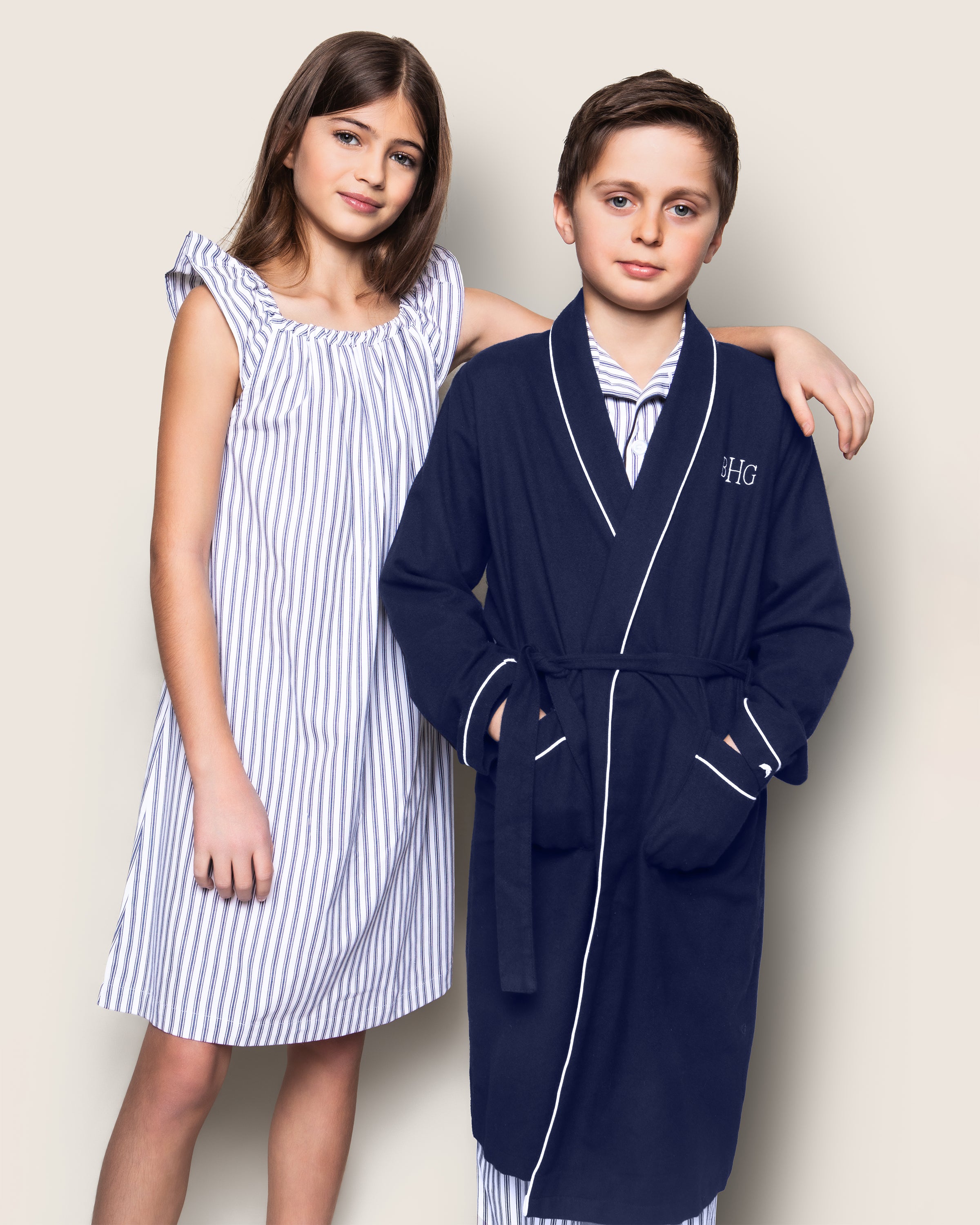 A girl in a striped dress stands next to a boy wrapped in Petite Plumes Kids Flannel Robe in Navy with White Piping, showcasing HG embroidery. Beneath, he wears flame-retardant striped pajamas. Both smile softly against a neutral background, highlighting their charming sleepwear.