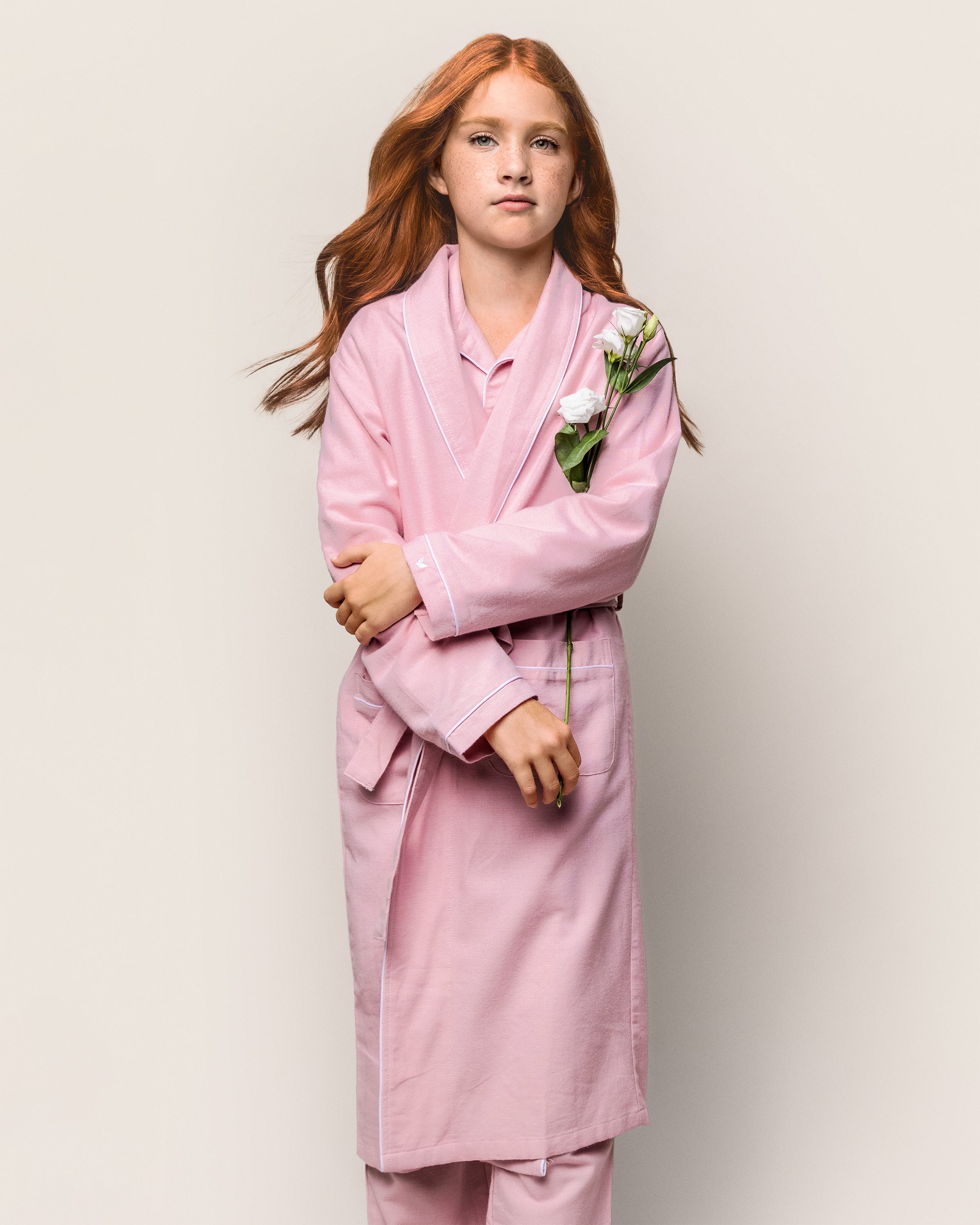 A young girl with long red hair stands against a plain background, wearing Petite Plumes Kids Flannel Robe in Pink and matching flame-retardant fiber pajamas. She holds white flowers across her chest, appearing calm and serene.