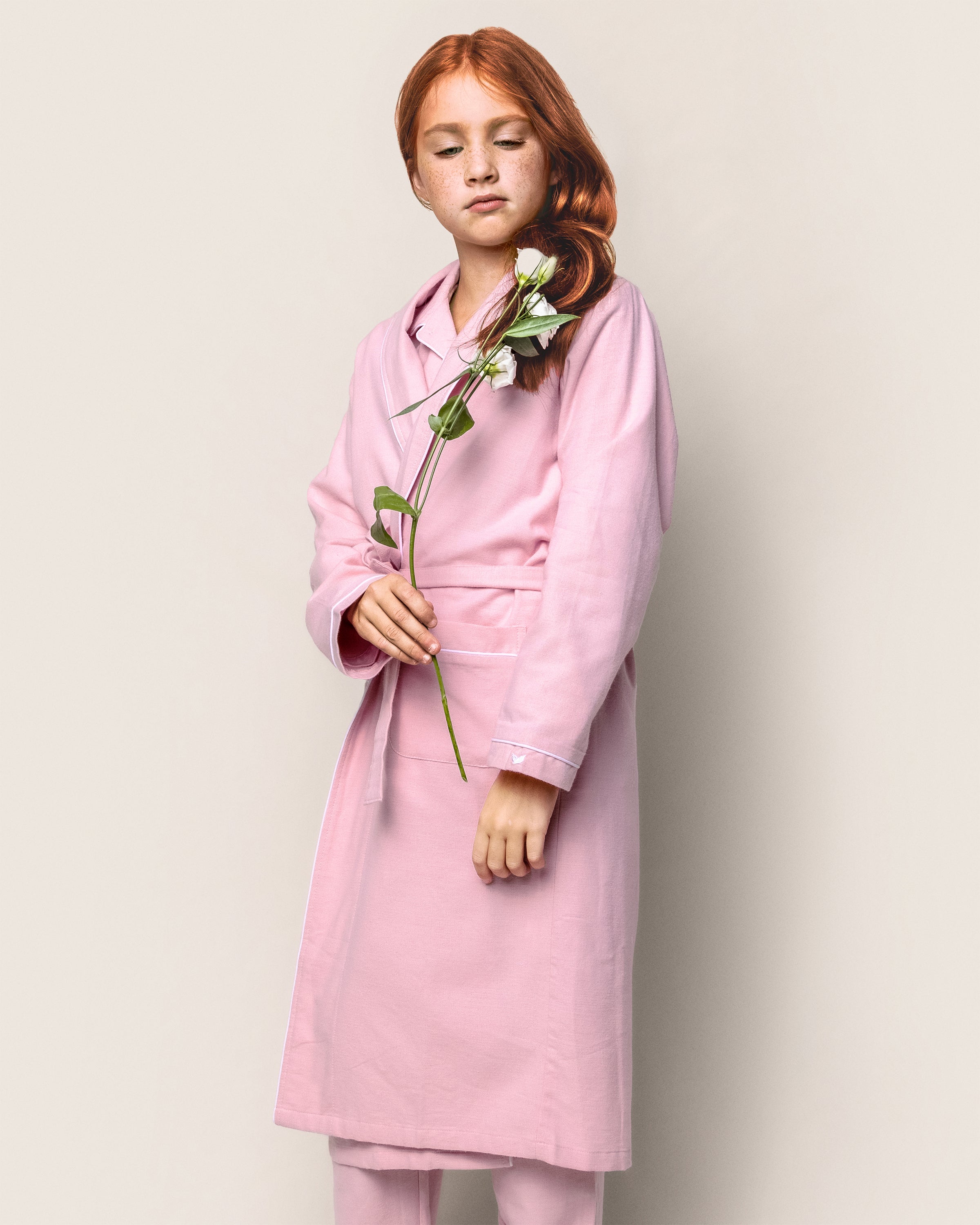 A person with long red hair stands against a plain background, wearing a Petite Plume Kids Flannel Robe in Pink. They hold a white flower gently in their hands, looking down thoughtfully.