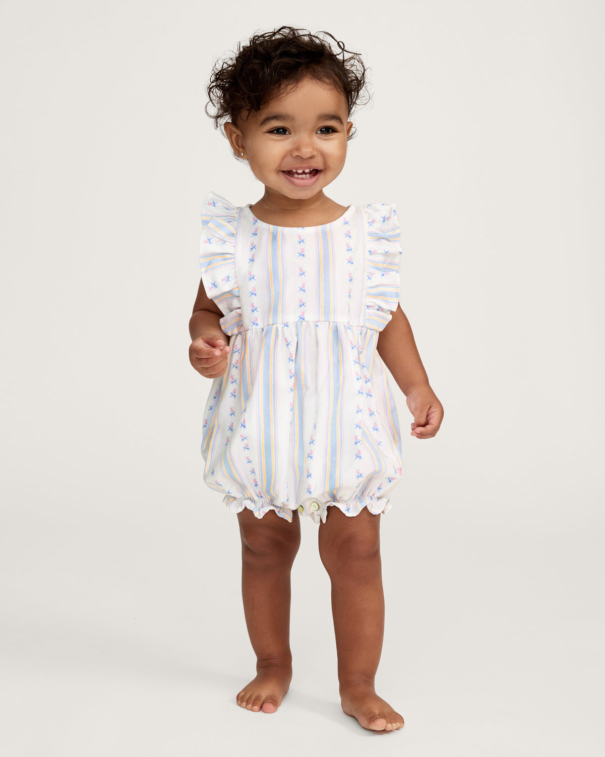 A smiling toddler with curly hair stands barefoot against a plain white background, wearing the Baby's Twill Ruffled Romper in Cottage Stripe—featuring pastel stripes, small floral patterns, and ruffled sleeves.