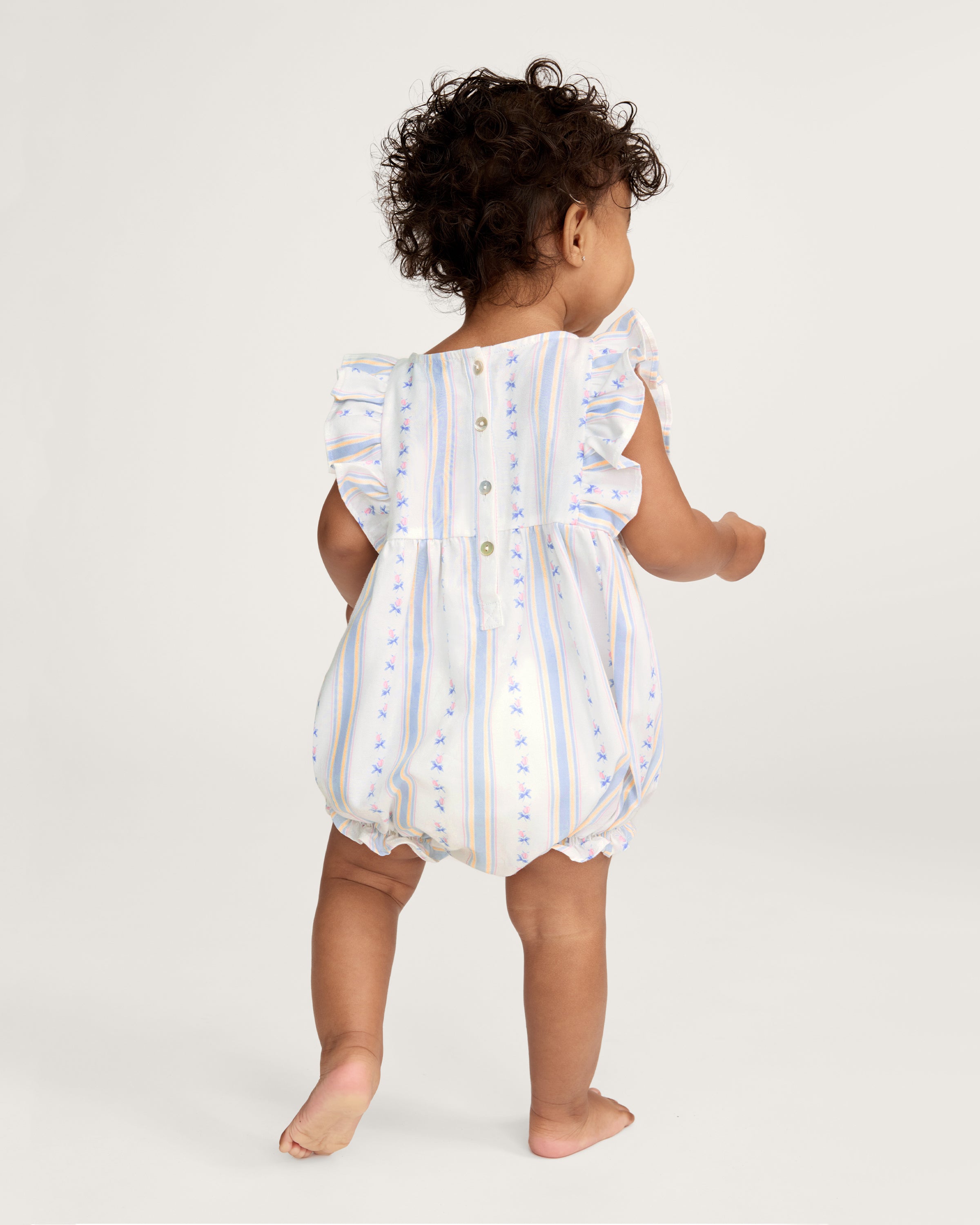 A smiling toddler with curly hair stands barefoot against a plain white background, wearing the Baby's Twill Ruffled Romper in Cottage Stripe—featuring pastel stripes, small floral patterns, and ruffled sleeves.