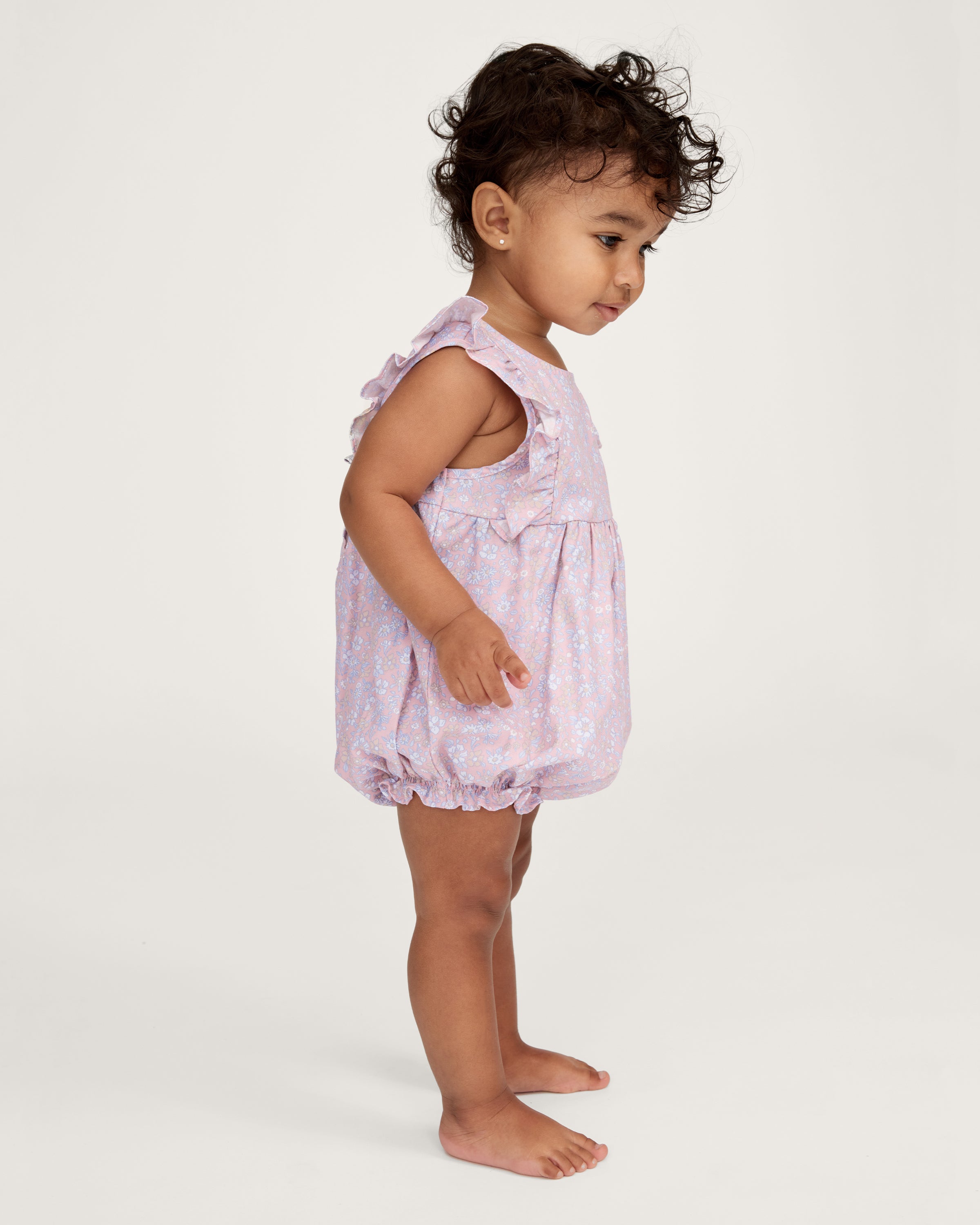 A curly-haired toddler stands barefoot, pointing left and looking that way, wearing the Baby's Twill Ruffled Romper in Fleur Blush against a plain light background.