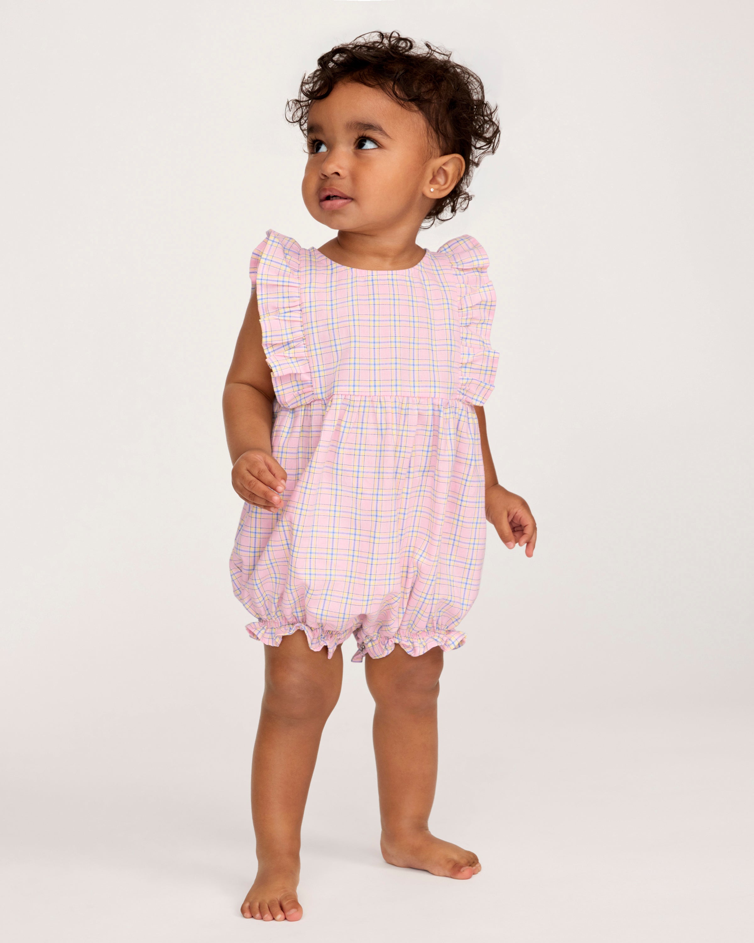 A toddler with curly hair wears the Baby's Twill Ruffled Romper in Peony Plaid, featuring ruffled sleeves, standing barefoot against a plain light background and looking slightly to the side.