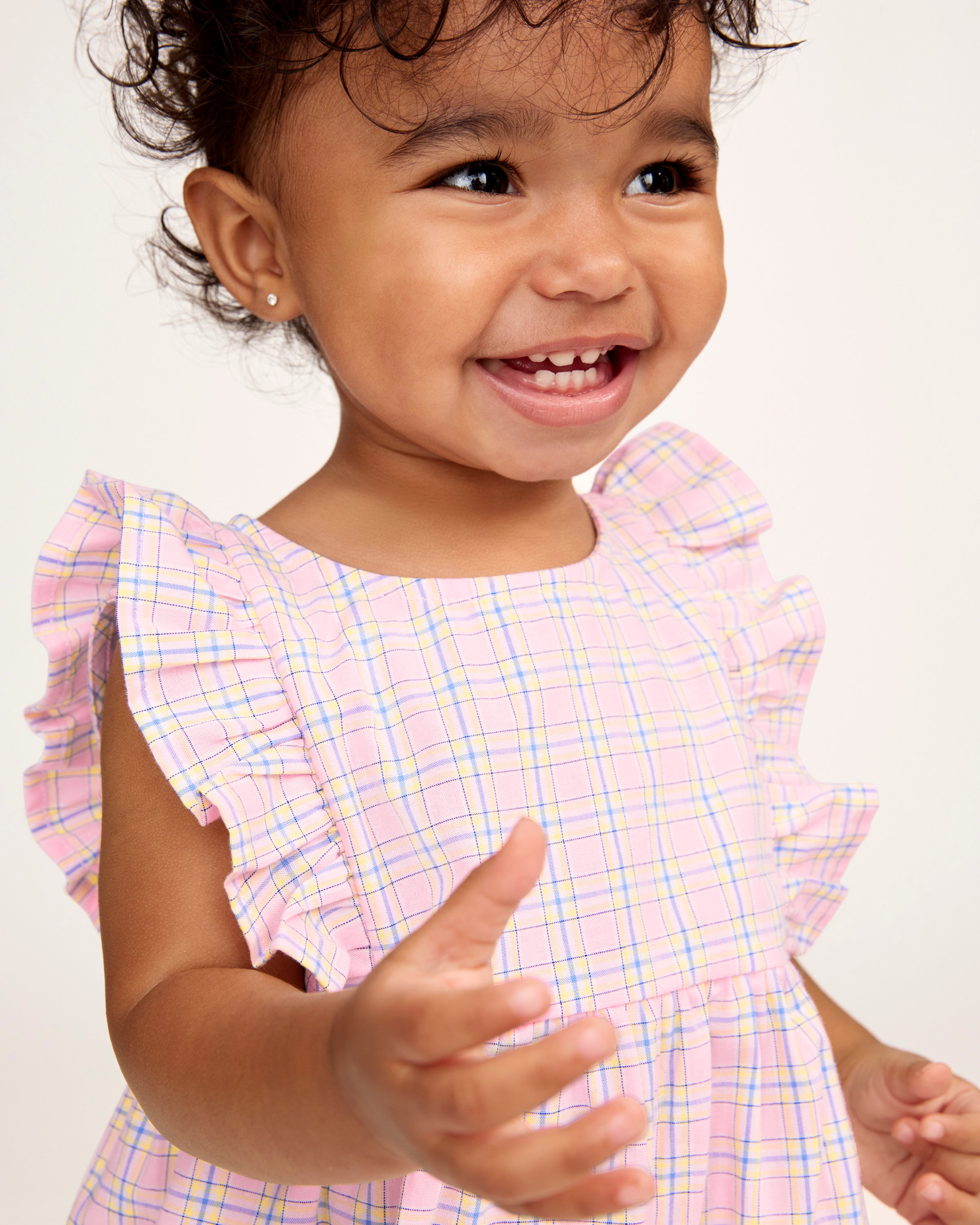 A toddler with curly hair wears the Baby's Twill Ruffled Romper in Peony Plaid, featuring ruffled sleeves, standing barefoot against a plain light background and looking slightly to the side.