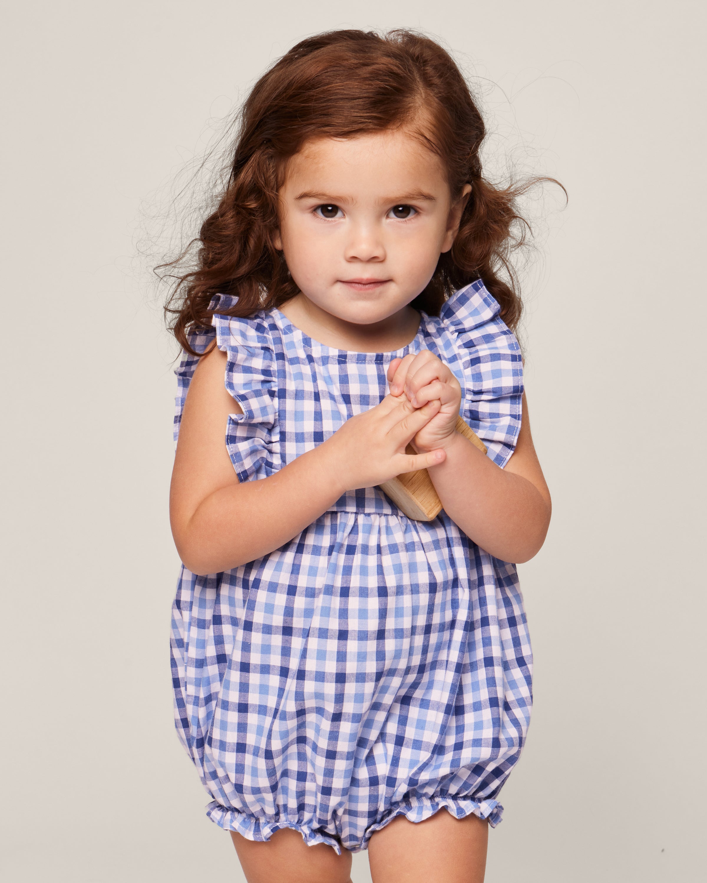 A toddler in Petite Plumes Babys Twill Ruffled Romper in Royal Blue Gingham stands against a plain background, showcasing its ruffled sleeves. She holds a wooden toy while looking forward with a slight smile.