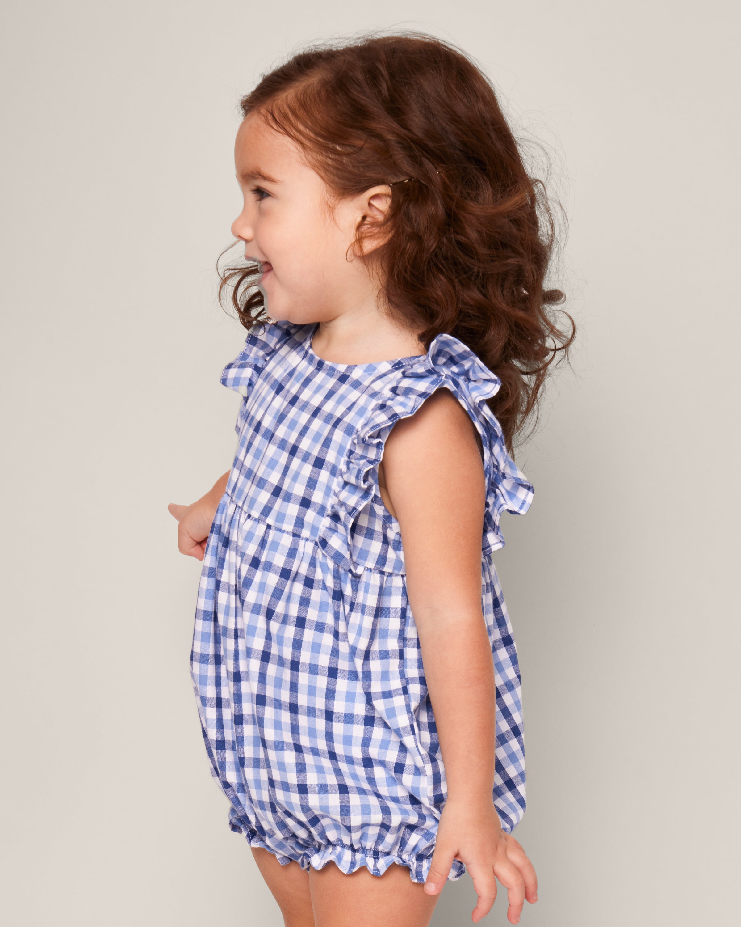 A toddler with curly brown hair stands sideways, smiling gently while wearing the Petite Plume Babys Twill Ruffled Romper in Royal Blue Gingham, set against a plain light grey background.