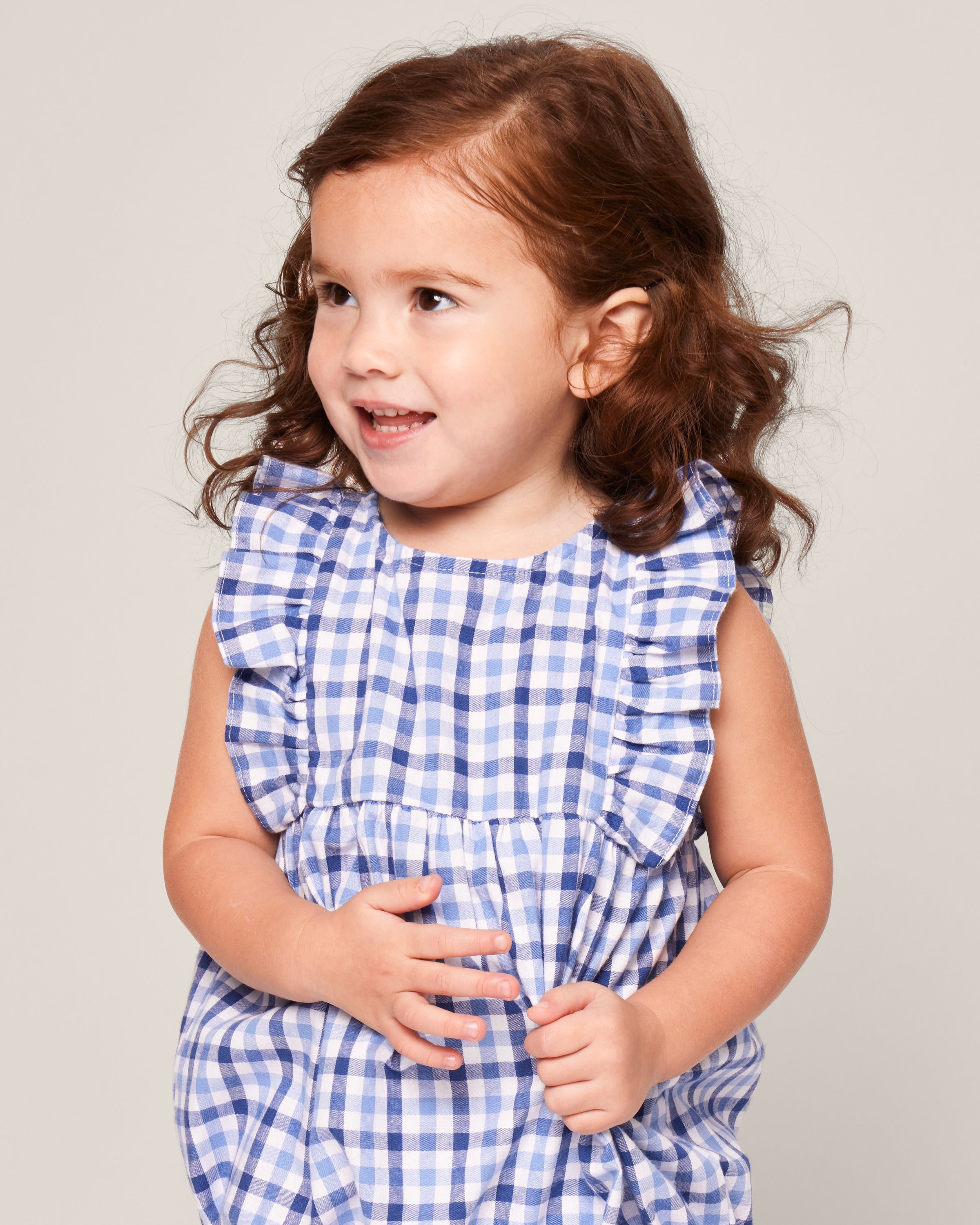A young child with wavy brown hair wears Petite Plumes Babys Twill Ruffled Romper in Royal Blue Gingham, smiling and looking sideways against a plain background.