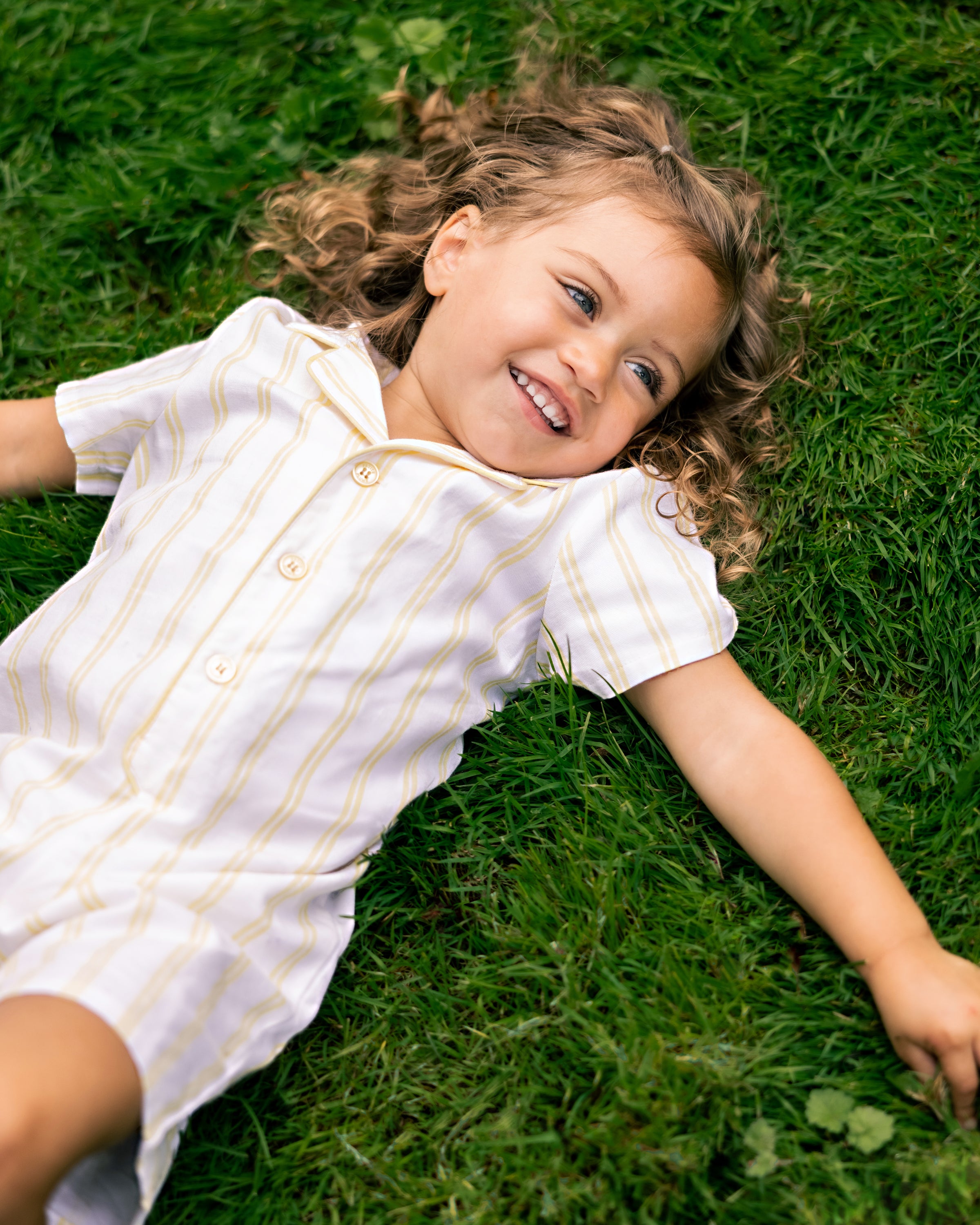 A young curly-haired child smiles while lying on green grass in a Babys Twill Summer Romper in Sunny Stripe by Petite Plume, enjoying a relaxed moment outdoors. The summer romper features soft yarn-dyed cotton, as the child gazes to the side, savoring the serene day.