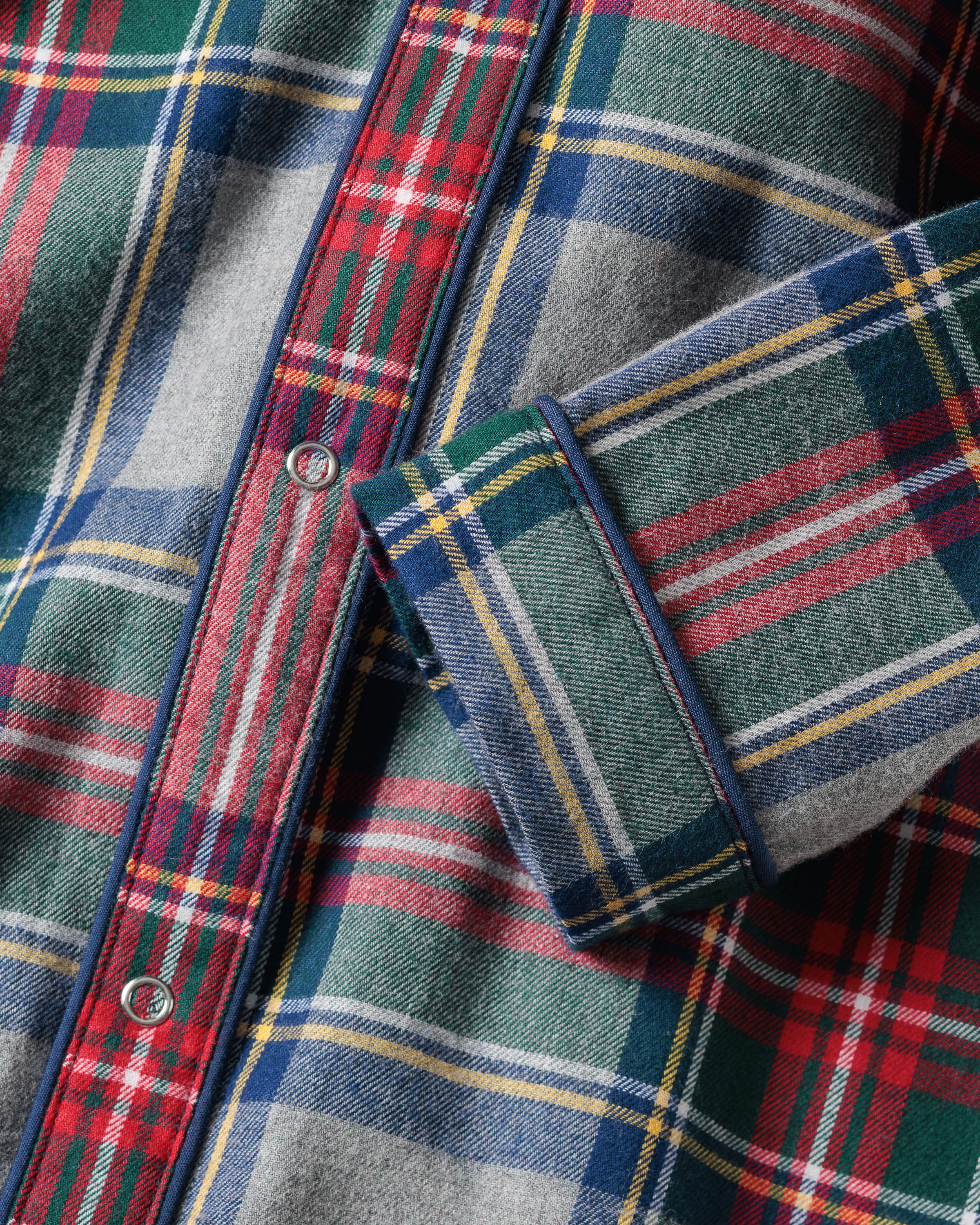 Close-up of Petite Plumes Babys Brushed Cotton Cambridge Romper in Westminster Tartan, showcasing red, blue, green, and yellow plaid. The image focuses on the front buttons and a partially folded sleeve, highlighting its detailed pattern inspired by Scottish tradition.