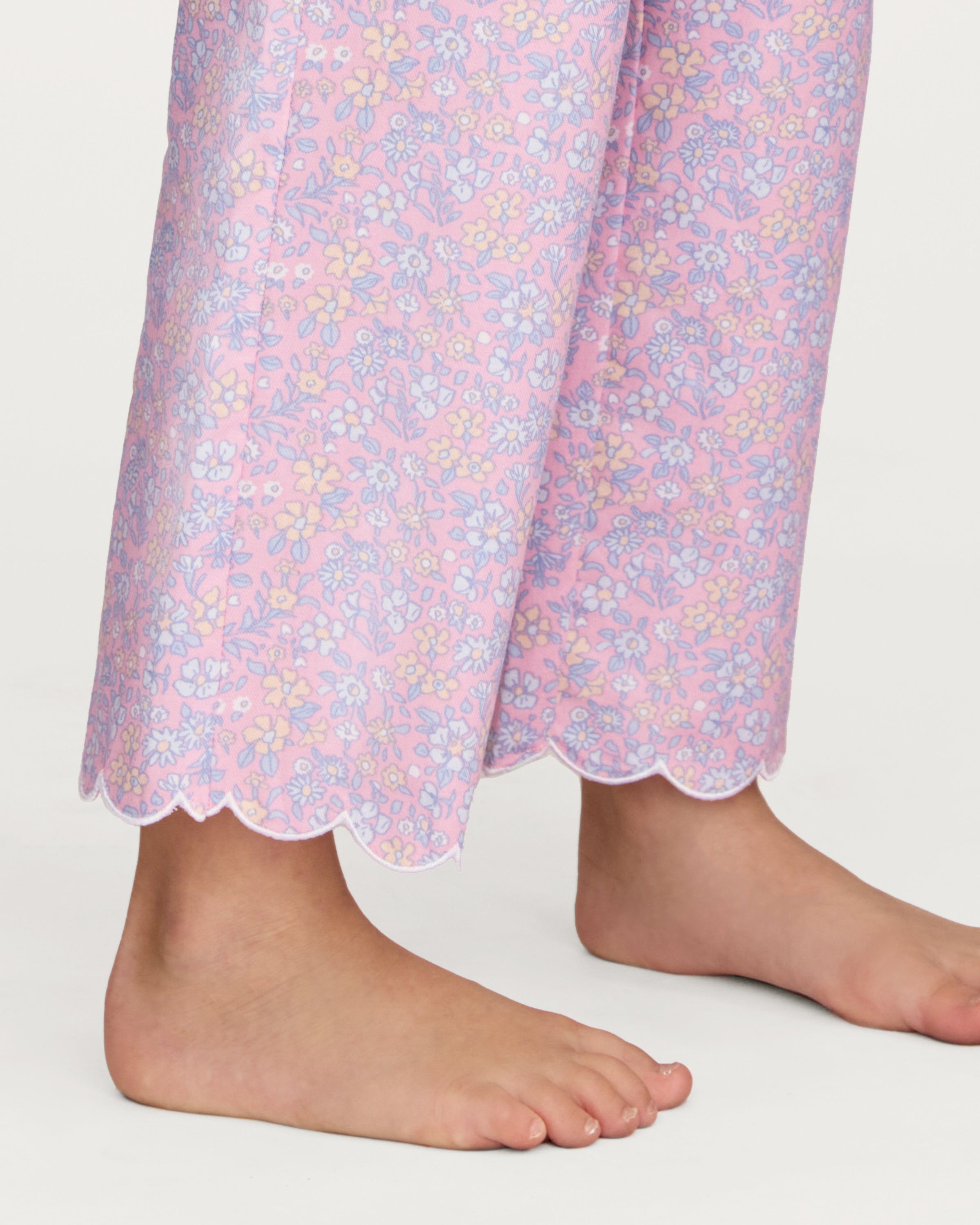 A young girl with wavy hair stands barefoot in the Kid's Twill Colette Pajama Set in Fleur Blush, featuring light pink floral fabric with a scalloped collar and pants, posing against a plain white background.