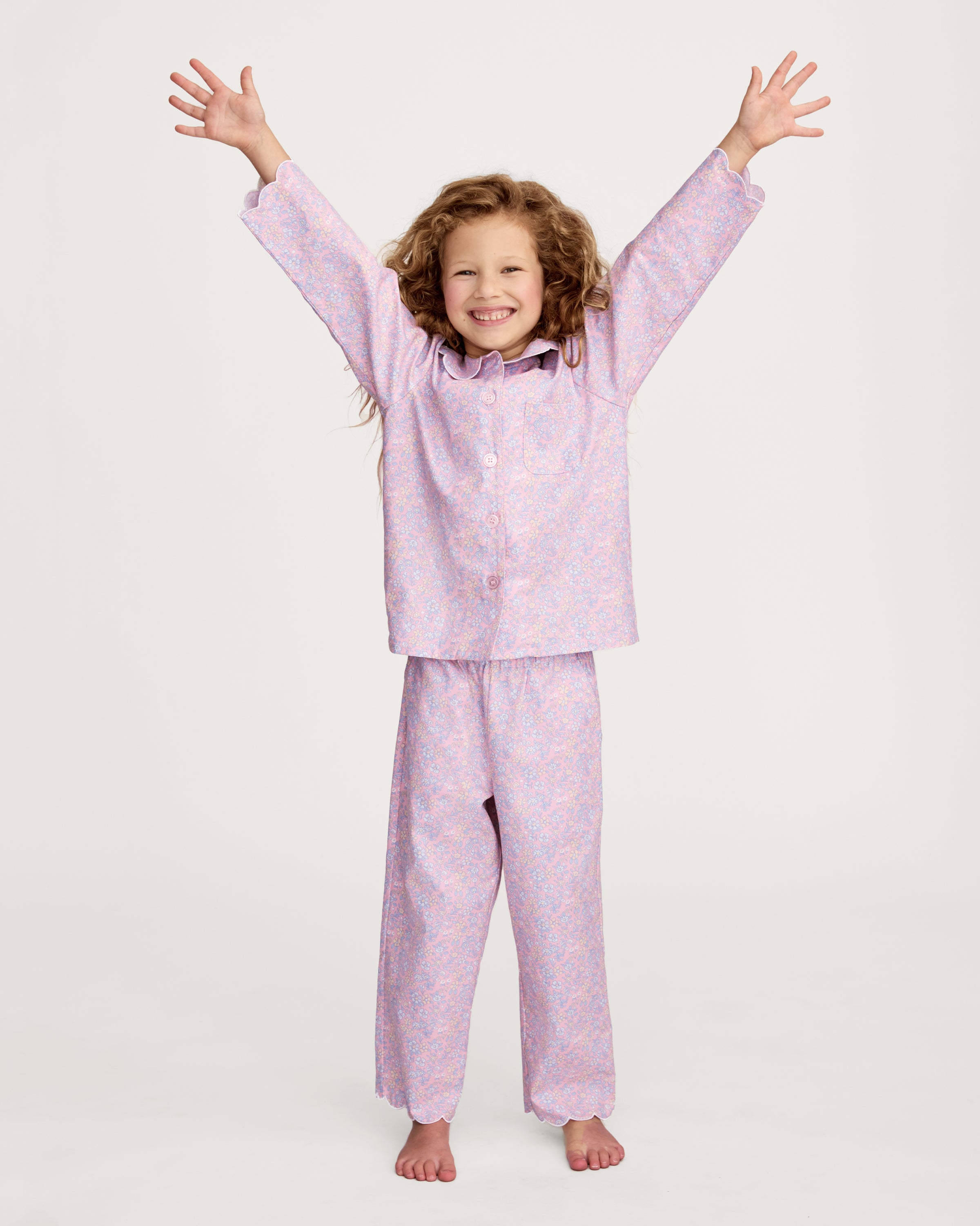 A young girl with wavy hair stands barefoot in the Kid's Twill Colette Pajama Set in Fleur Blush, featuring light pink floral fabric with a scalloped collar and pants, posing against a plain white background.