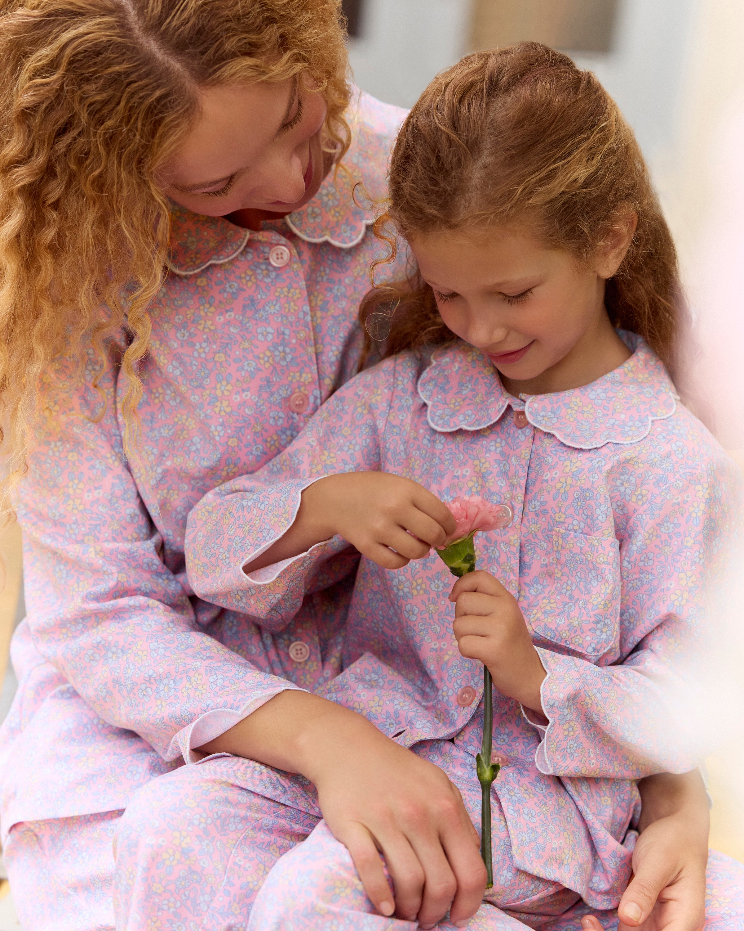 A young girl with wavy hair stands barefoot in the Kid's Twill Colette Pajama Set in Fleur Blush, featuring light pink floral fabric with a scalloped collar and pants, posing against a plain white background.