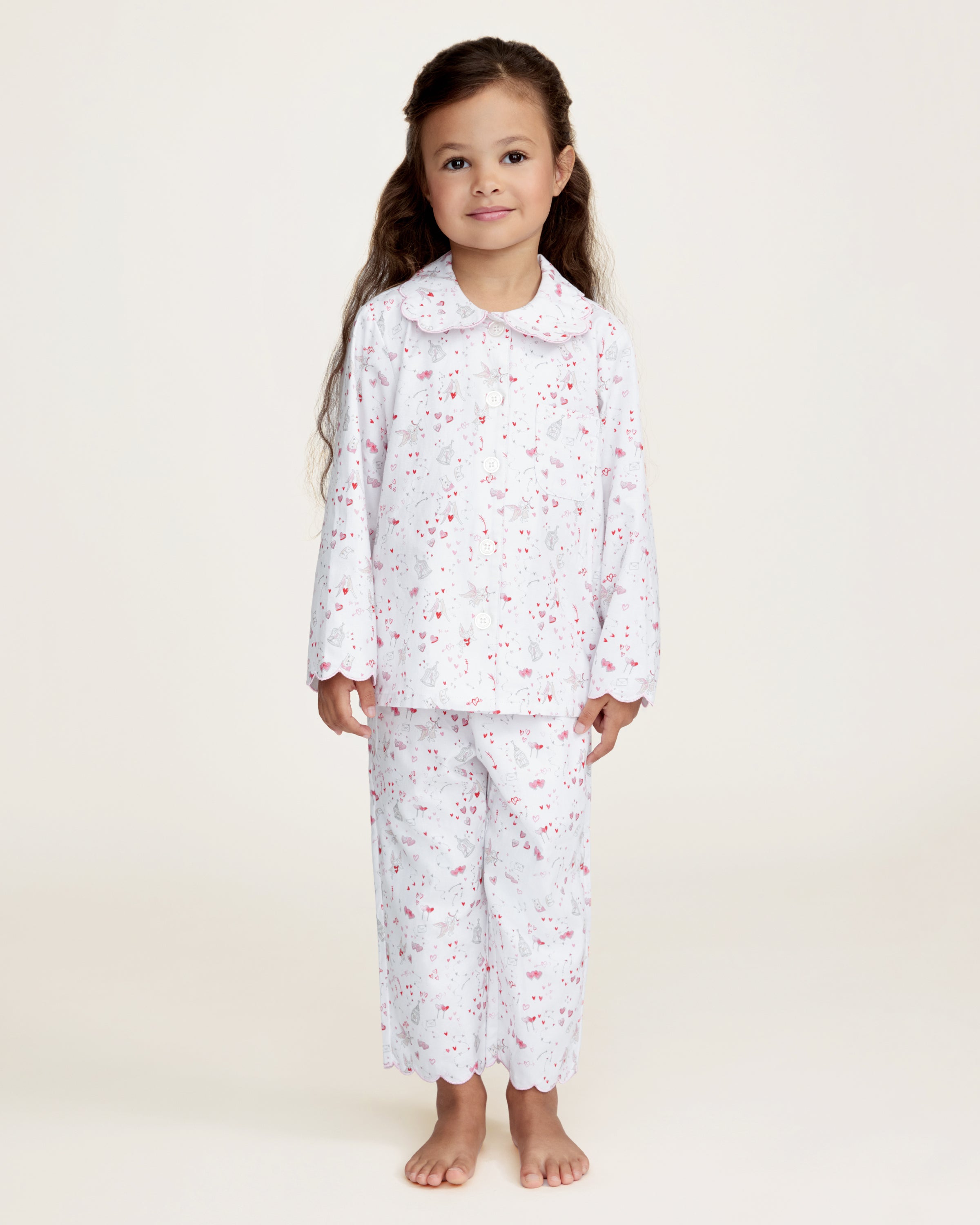 The Kid's Twill Colette Pajama Set in Love Song features white pajamas with a scalloped collar and cuffs, adorned with pink and red heart and dot patterns. The set includes a long-sleeved button-up top and matching pants.