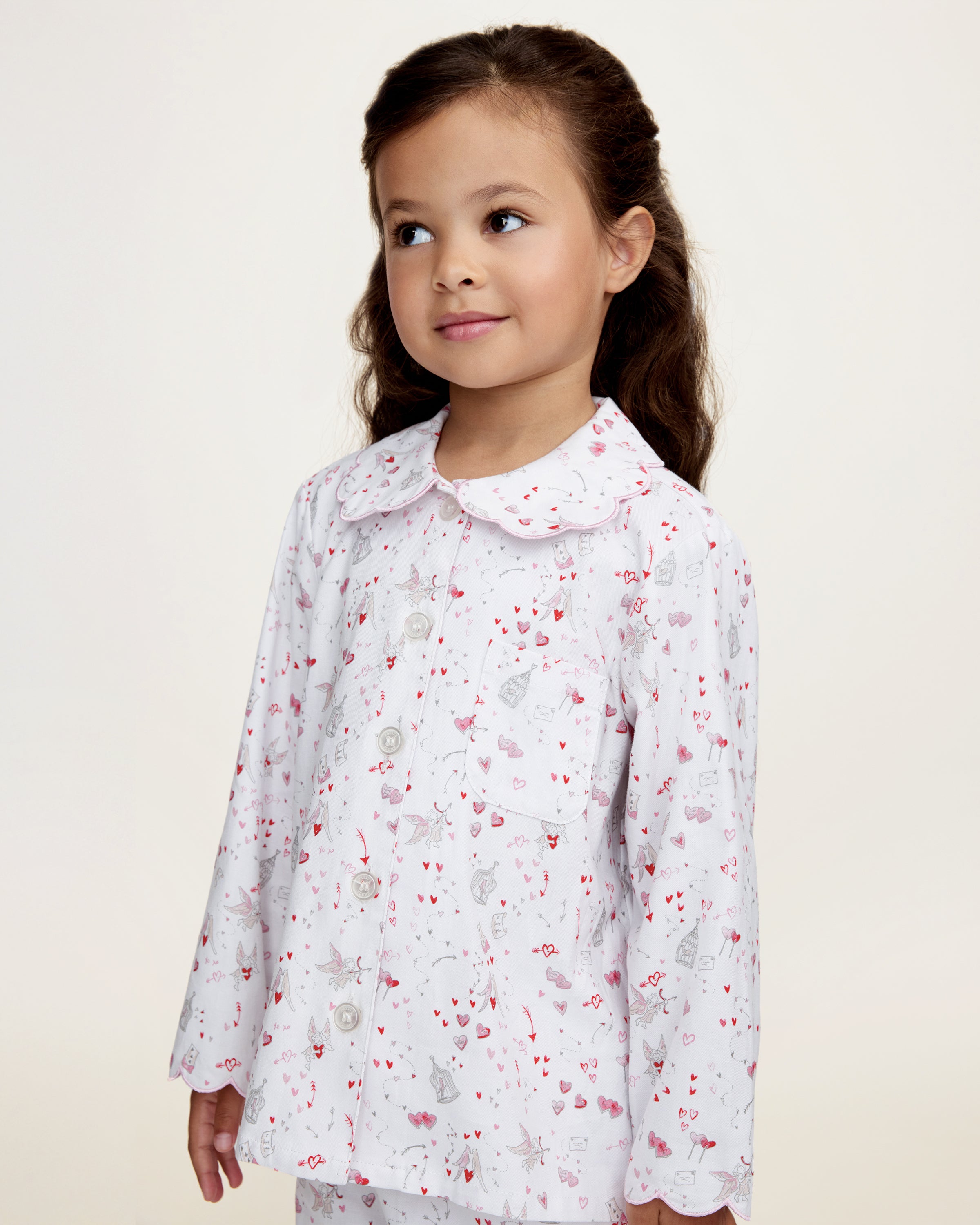 The Kid's Twill Colette Pajama Set in Love Song features white pajamas with a scalloped collar and cuffs, adorned with pink and red heart and dot patterns. The set includes a long-sleeved button-up top and matching pants.