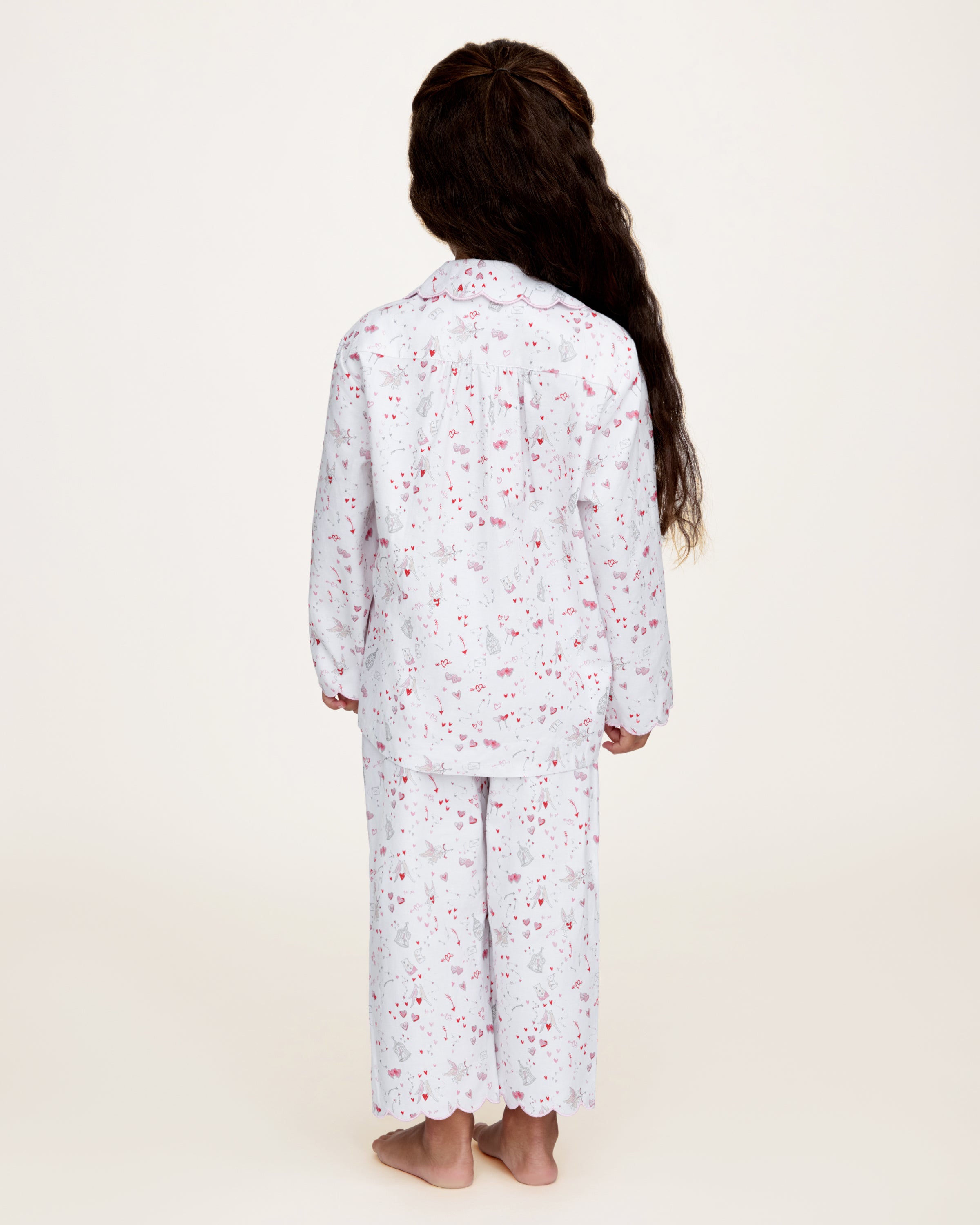 The Kid's Twill Colette Pajama Set in Love Song features white pajamas with a scalloped collar and cuffs, adorned with pink and red heart and dot patterns. The set includes a long-sleeved button-up top and matching pants.