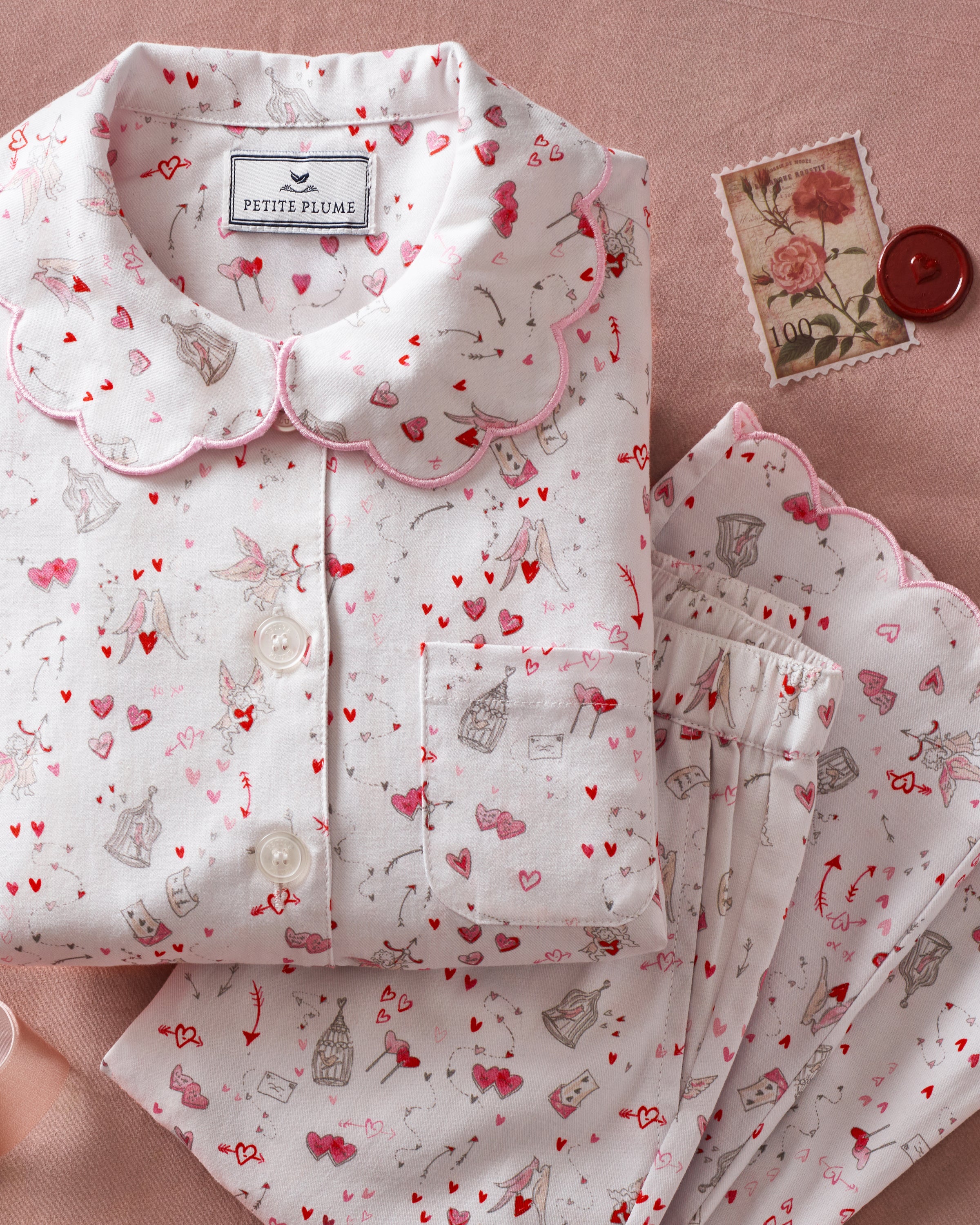 The Kid's Twill Colette Pajama Set in Love Song features white pajamas with a scalloped collar and cuffs, adorned with pink and red heart and dot patterns. The set includes a long-sleeved button-up top and matching pants.
