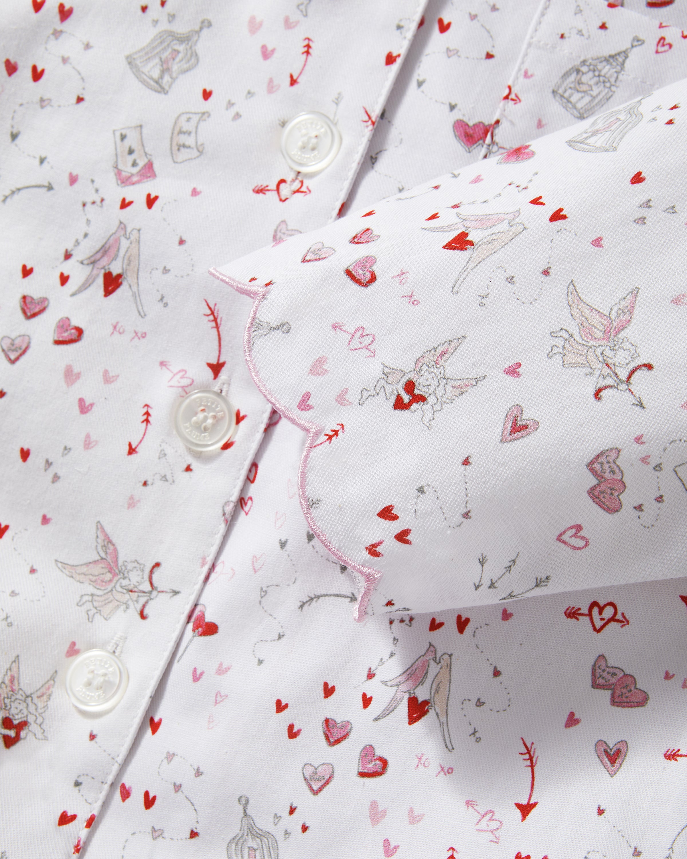 The Kid's Twill Colette Pajama Set in Love Song features white pajamas with a scalloped collar and cuffs, adorned with pink and red heart and dot patterns. The set includes a long-sleeved button-up top and matching pants.