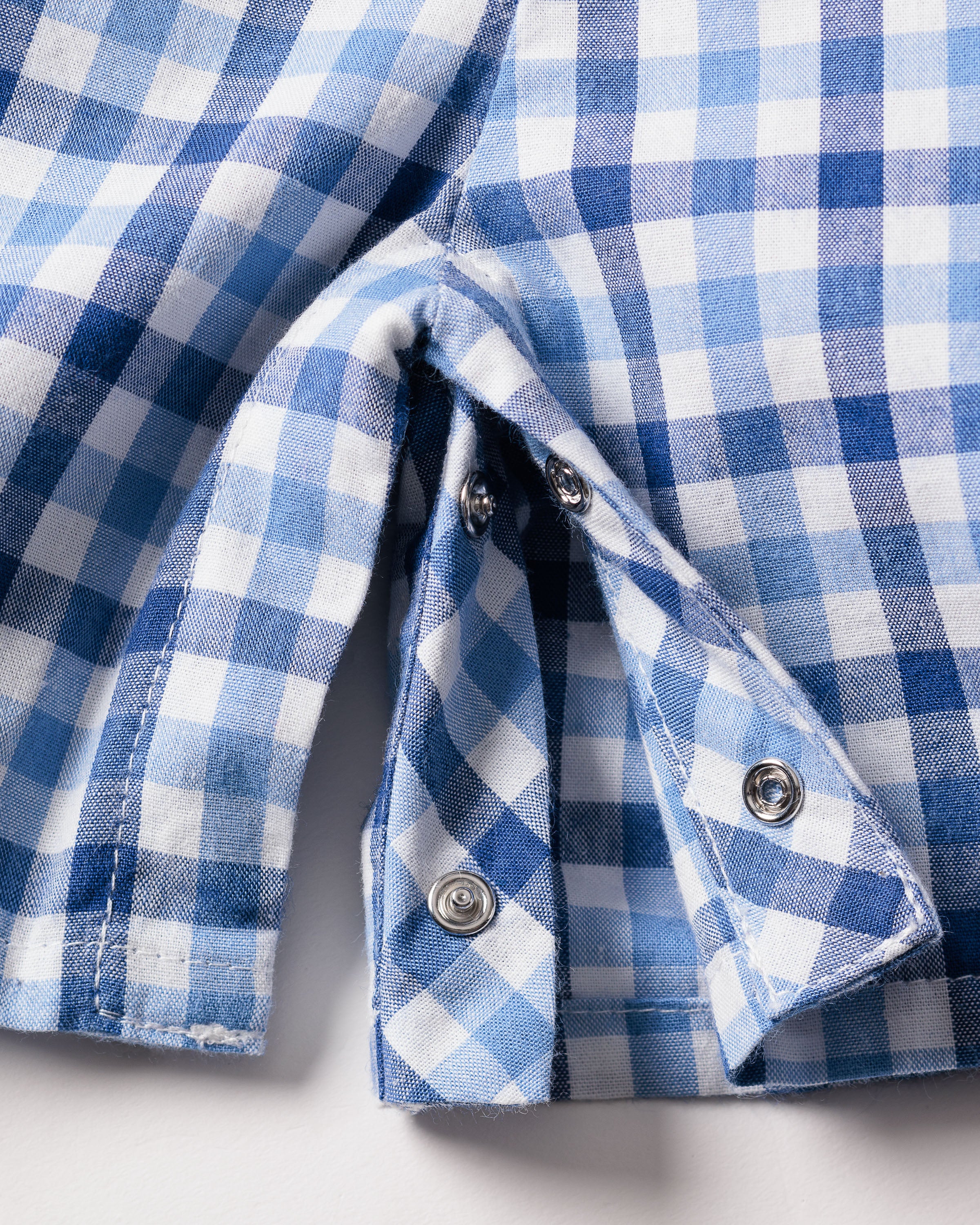 Close-up of Petite Plumes Babys Twill Summer Romper in Royal Blue Gingham, highlighting the metal snap buttons. The yarn-dyed cotton fabric is slightly folded to reveal detailed stitching and button features.