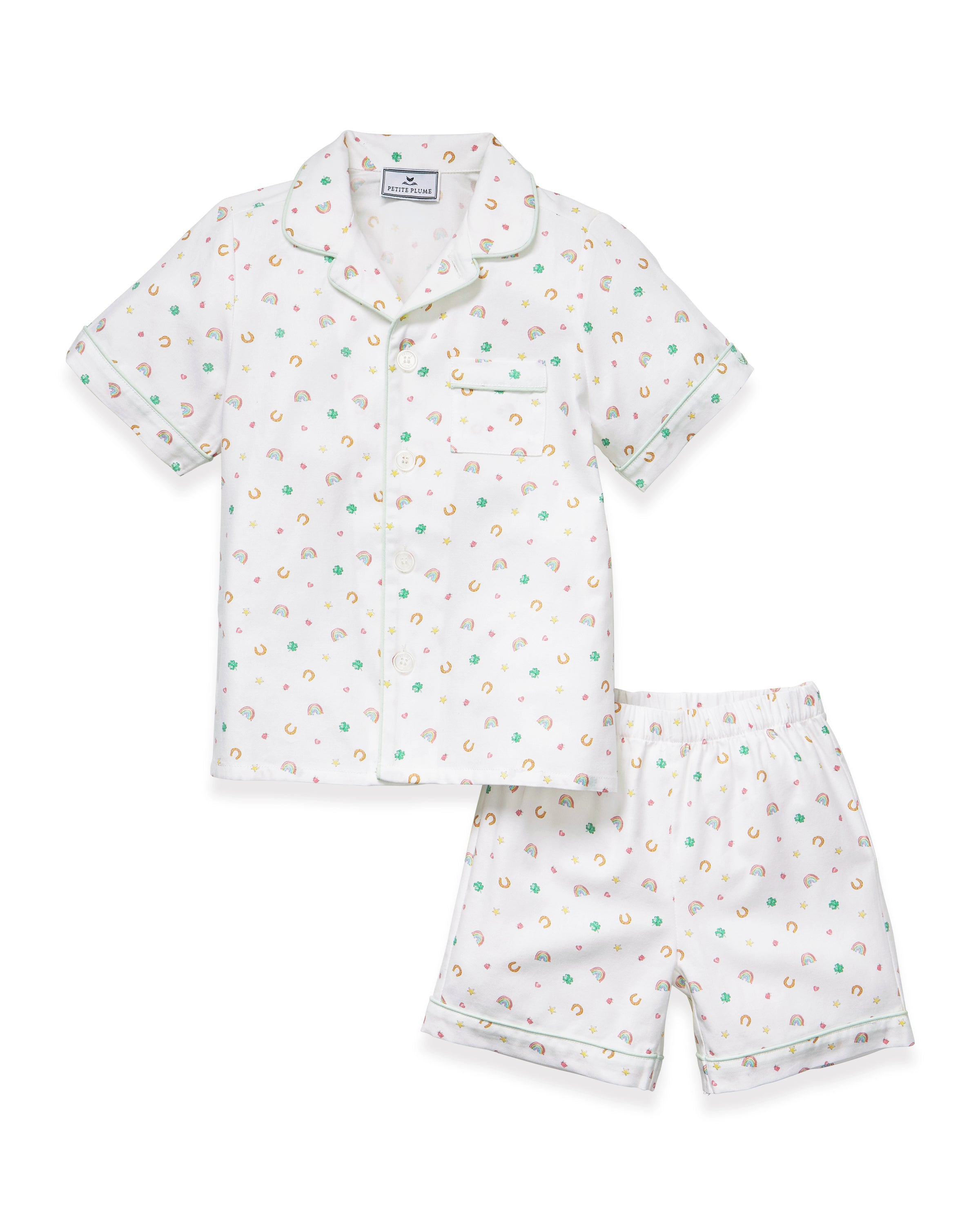 A close-up of a child in the Kid's Twill Pajama Short Set in Good Luck Charms, featuring white fabric with light green trim and pastel rainbows, horseshoes, four-leaf clovers, and stars. Only the upper torso is shown.