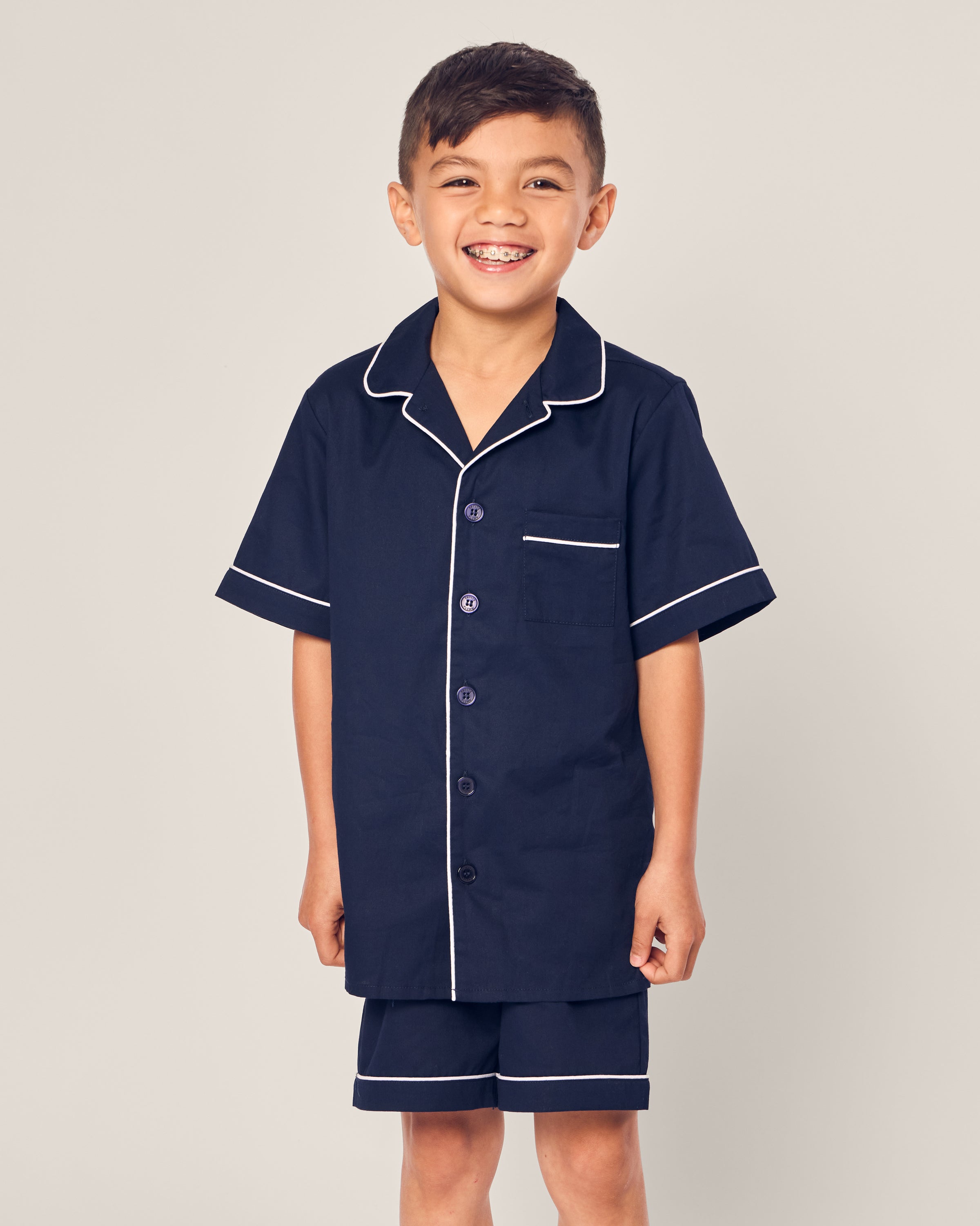 A smiling child wears the Kids Twill Short Sleeve Short Set in Navy by Petite Plume, crafted from luxurious sleepwear fabric. The set, with white piping, includes a short-sleeved button-up shirt and matching shorts. A plain beige background enhances its timeless elegance.