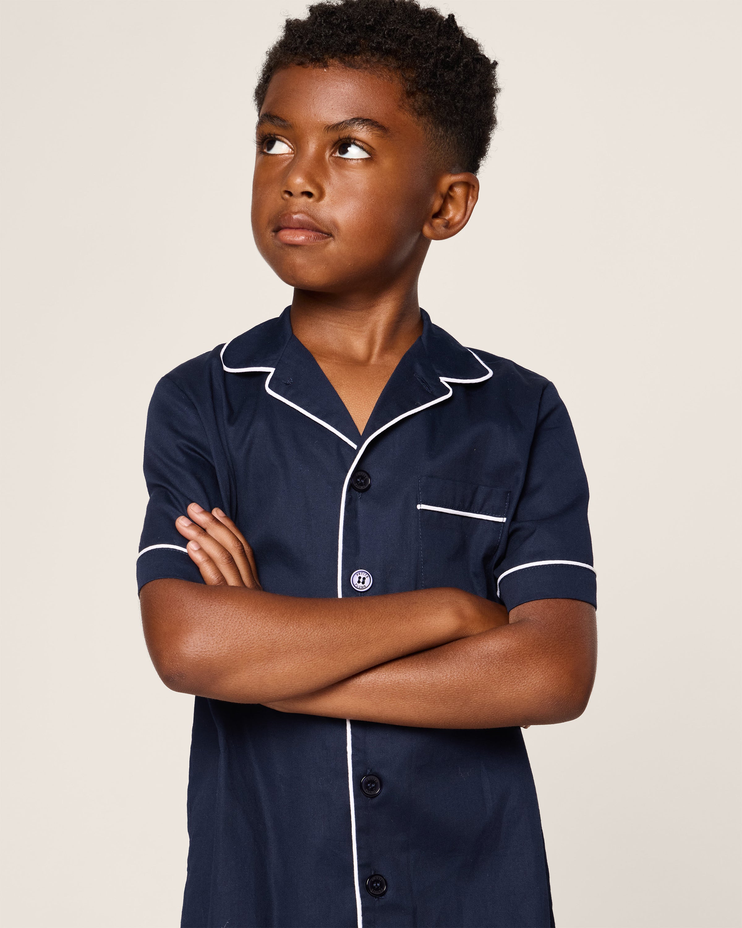 A child with short curly hair stands with arms crossed, looking up to the side, wearing the Petite Plume Kids Twill Short Sleeve Short Set in Navy. This outfit features white piping details on the collar and sleeves against a plain, light background.