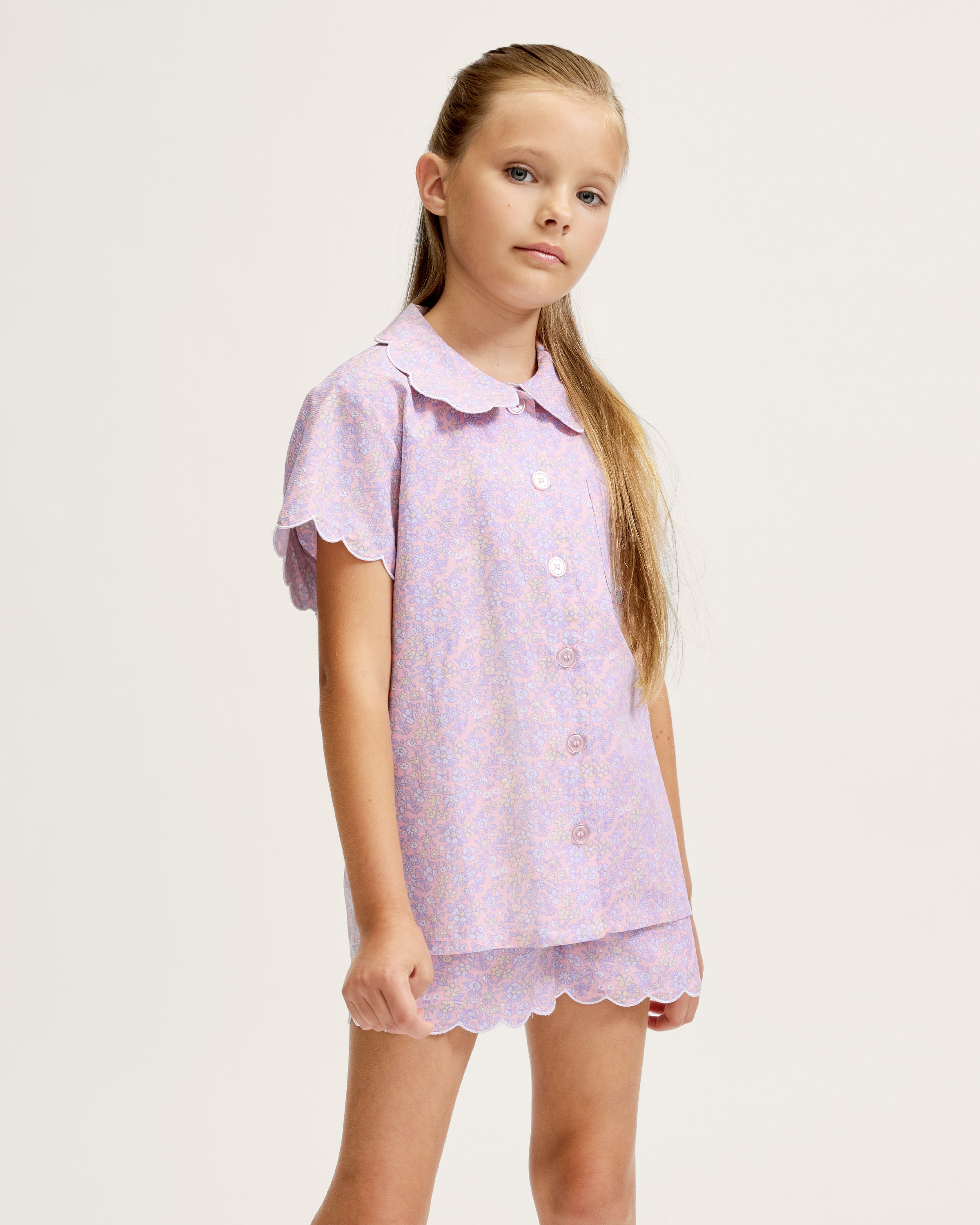A young girl with long blonde hair stands facing forward, wearing the Kid's Twill Colette Short Set in Fleur Blush—an adorable pastel purple and pink pajama set with scalloped edges—against a plain light background.