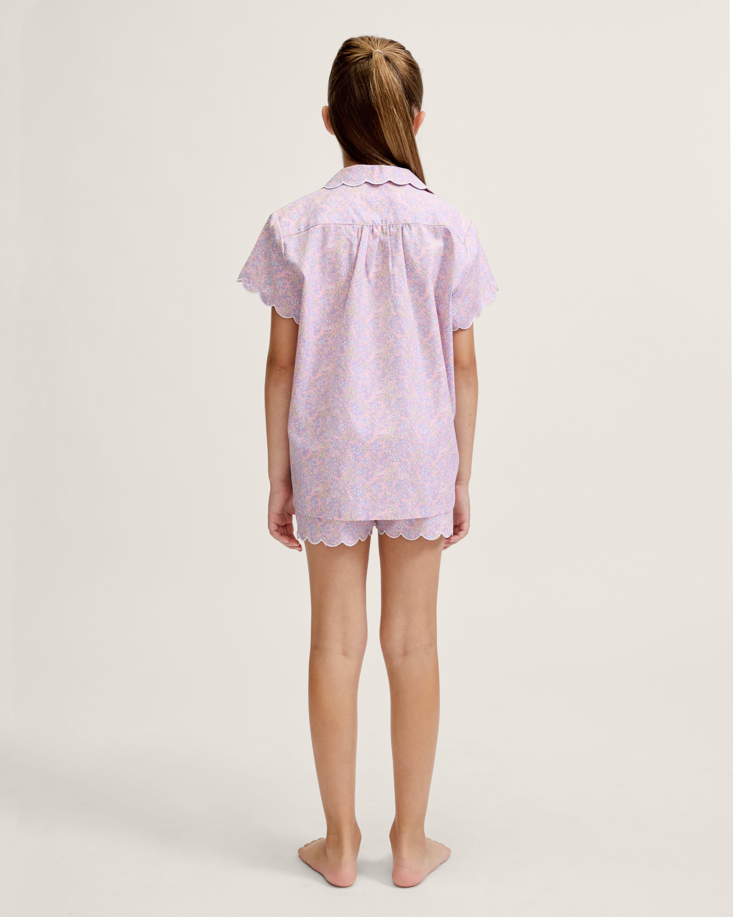A young girl with long blonde hair stands facing forward, wearing the Kid's Twill Colette Short Set in Fleur Blush—an adorable pastel purple and pink pajama set with scalloped edges—against a plain light background.