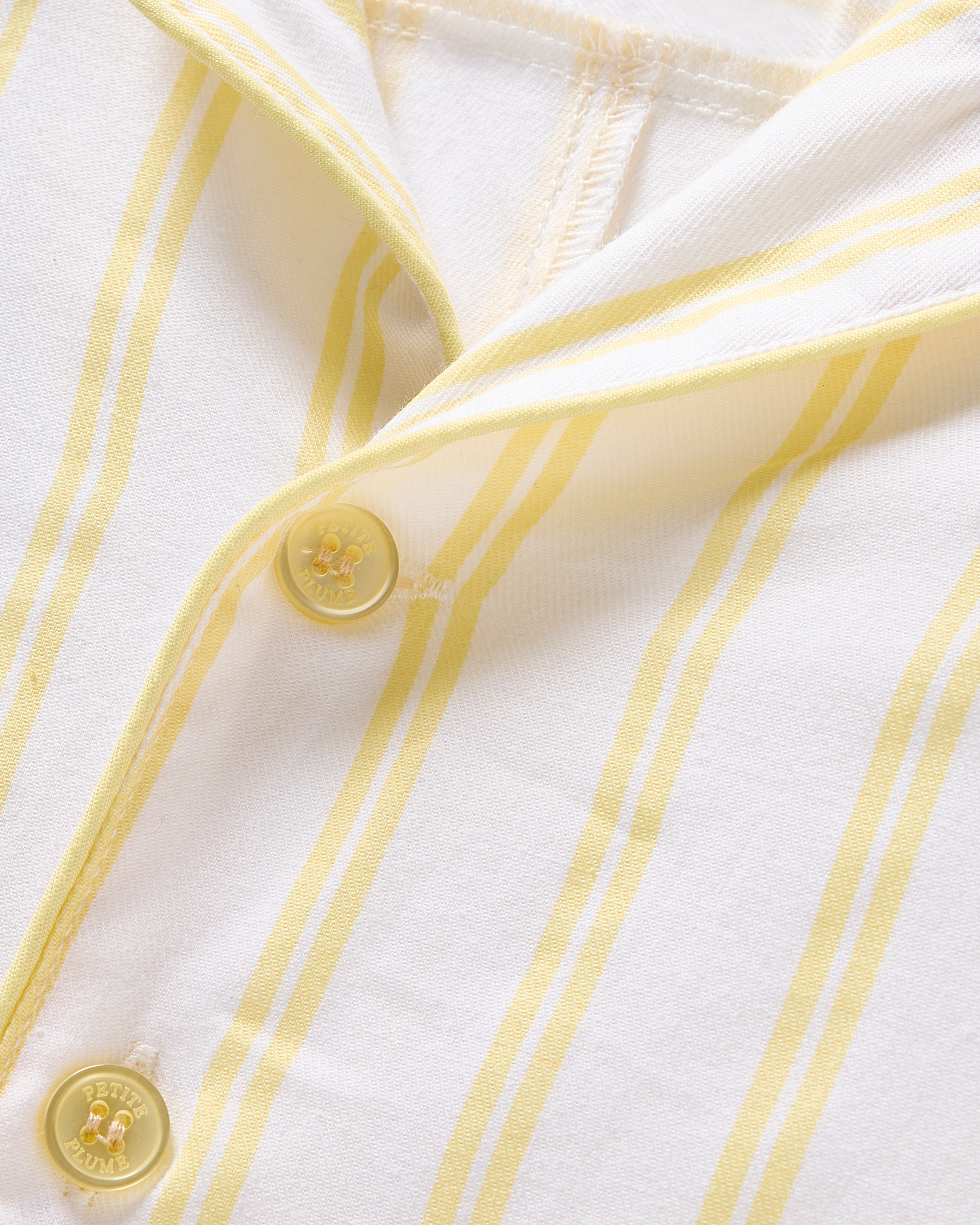 Close-up of Petite Plumes Kids Twill Pajama in Sunny Stripe, showcasing yellow and white yarn-dyed cotton with a buttonhole, two yellow buttons, and part of a collar visible. The fabrics texture and sewing details are clearly shown.
