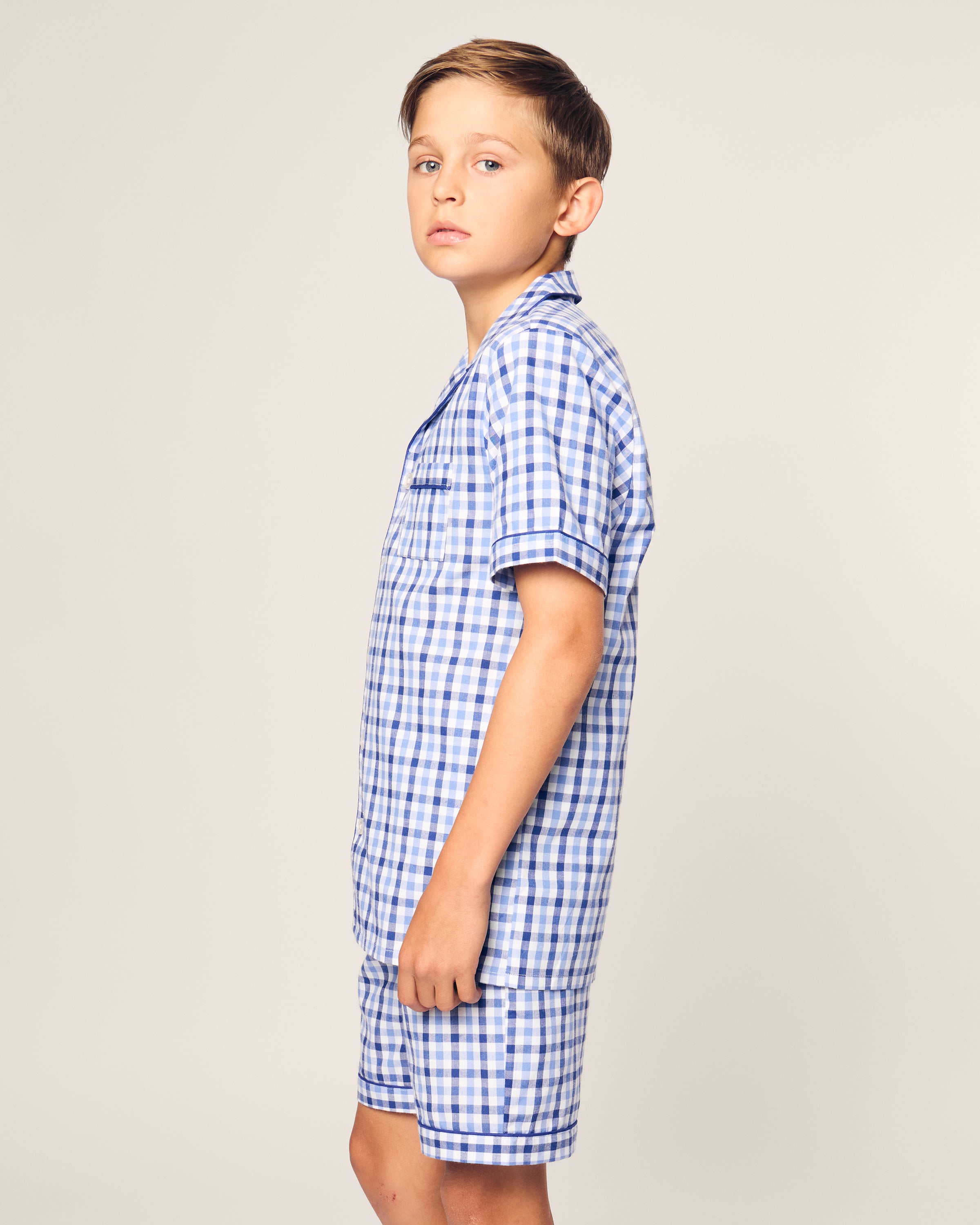 A young boy stands sideways, looking at the camera in a Kids Twill Pajama Short Set by Petite Plume. He wears a Royal Blue Gingham yarn-dyed cotton shirt and matching shorts against a plain light-colored background.