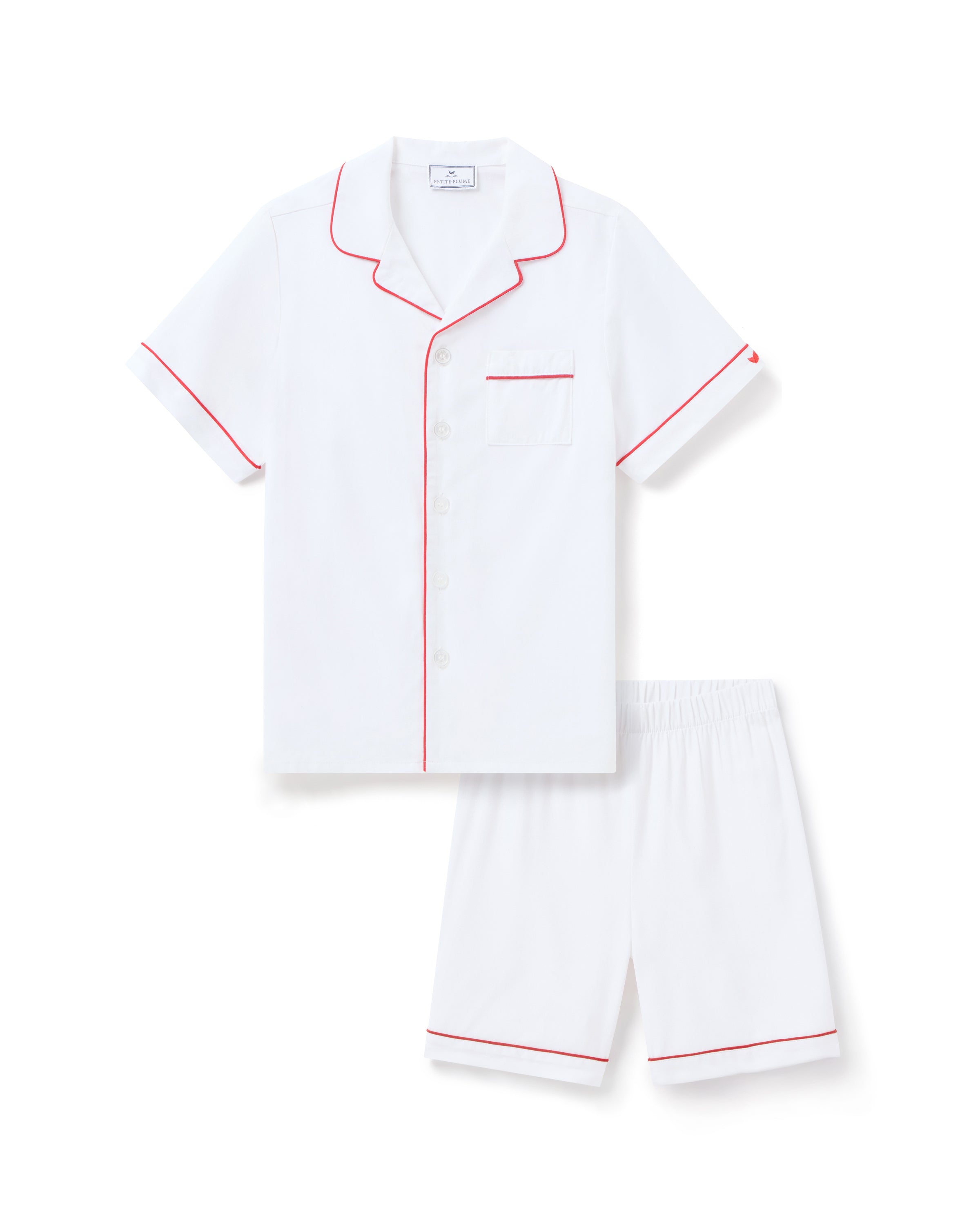 The Kids White Twill Short Set with Red Piping by Petite Plume, made from yarn-dyed cotton, includes a short-sleeve button-up shirt with a pocket and matching shorts, displayed on a plain background.