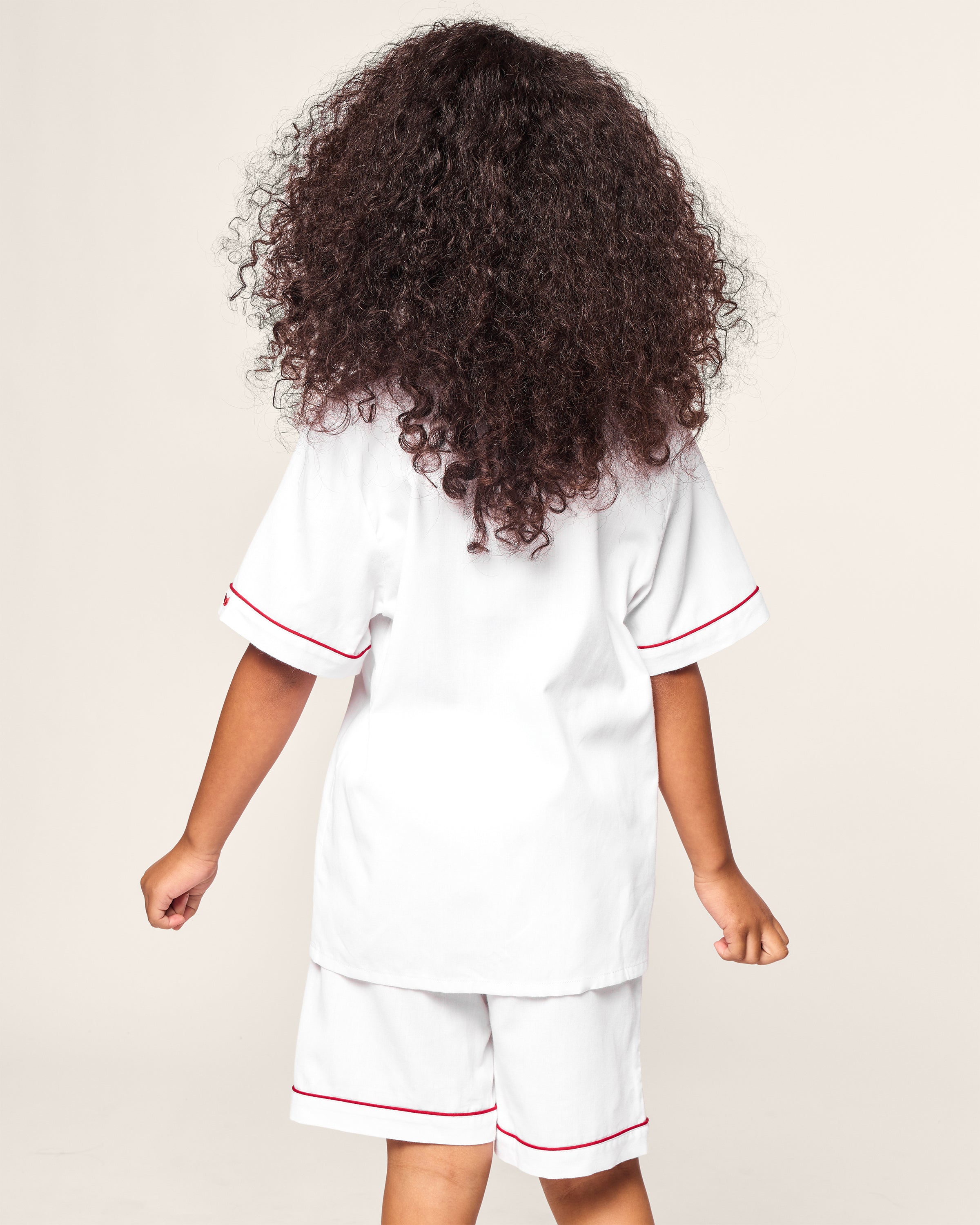 A child with curly hair is wearing Petite Plumes Kids White Twill Short Set with Red Piping, facing away.