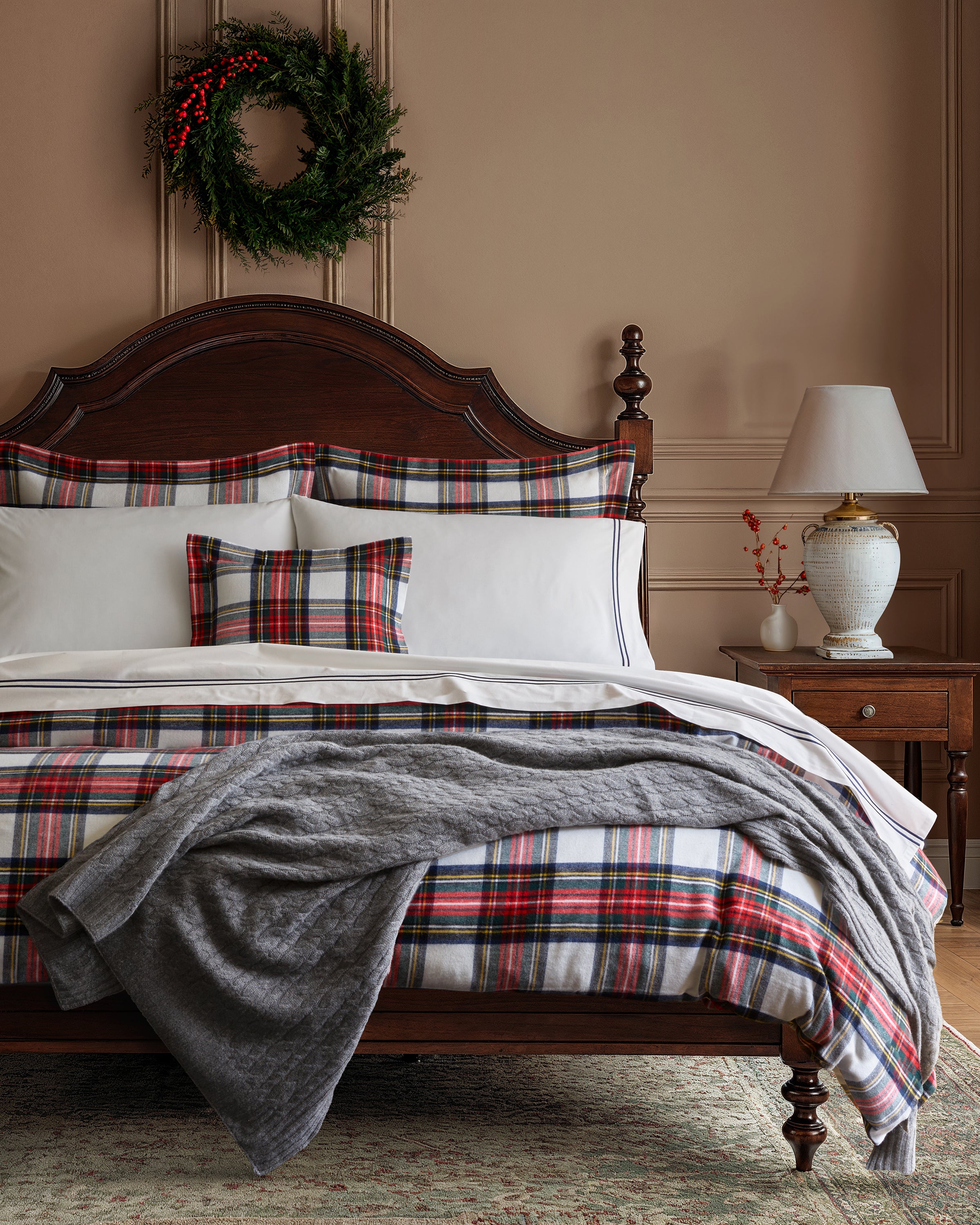A cozy bedroom with a wooden bed adorned in red, green, and white plaid bedding. A Petite Plume Cashmere Cable Knit Throw in Dark Grey is draped over the edge. The nightstand with a lamp and vase suits decor lovers. A green wreath hangs on the beige wall above the bed, and a textured rug covers the floor.