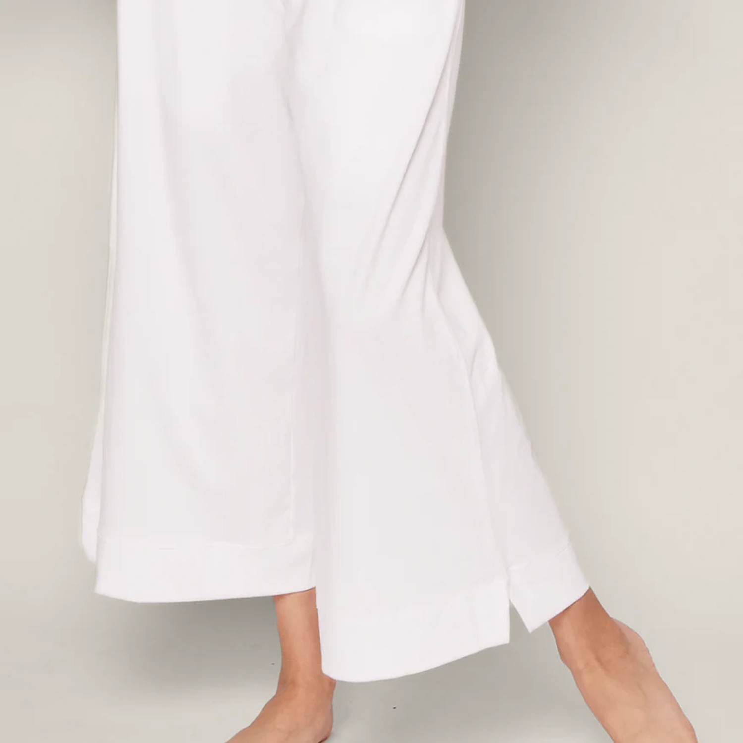 A person wearing loose-fitting white pants stands barefoot on a light background, with one foot slightly in front of the other.