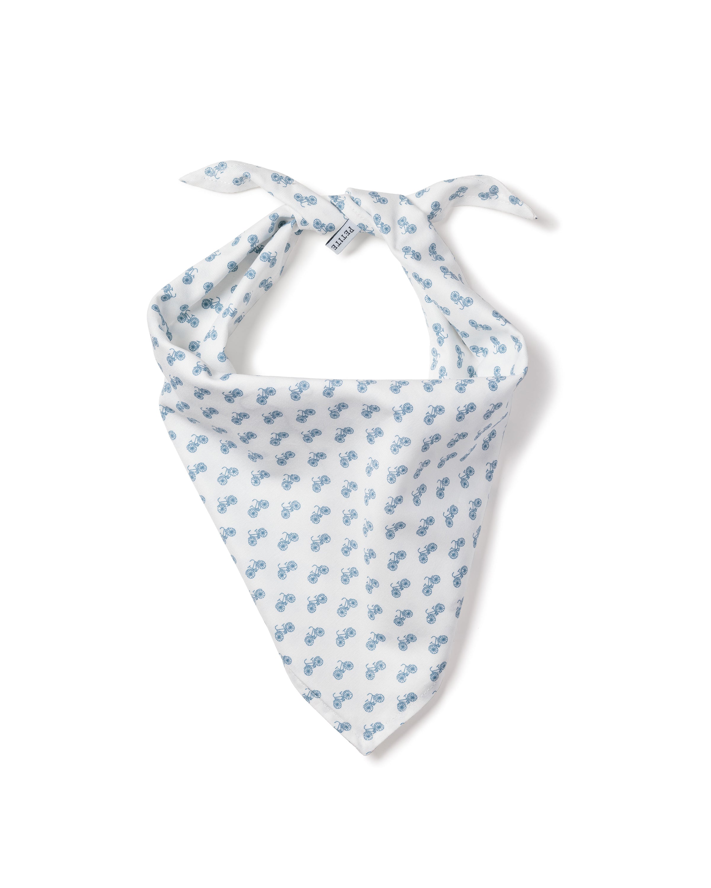 A fluffy white dog with a friendly expression lies on a wooden floor, wearing the charming Dog Twill Bandana in Bicyclette by Petite Plume. Its pointed ears and slightly open mouth reveal its tongue, enhancing the allure of this delightful dog accessory.