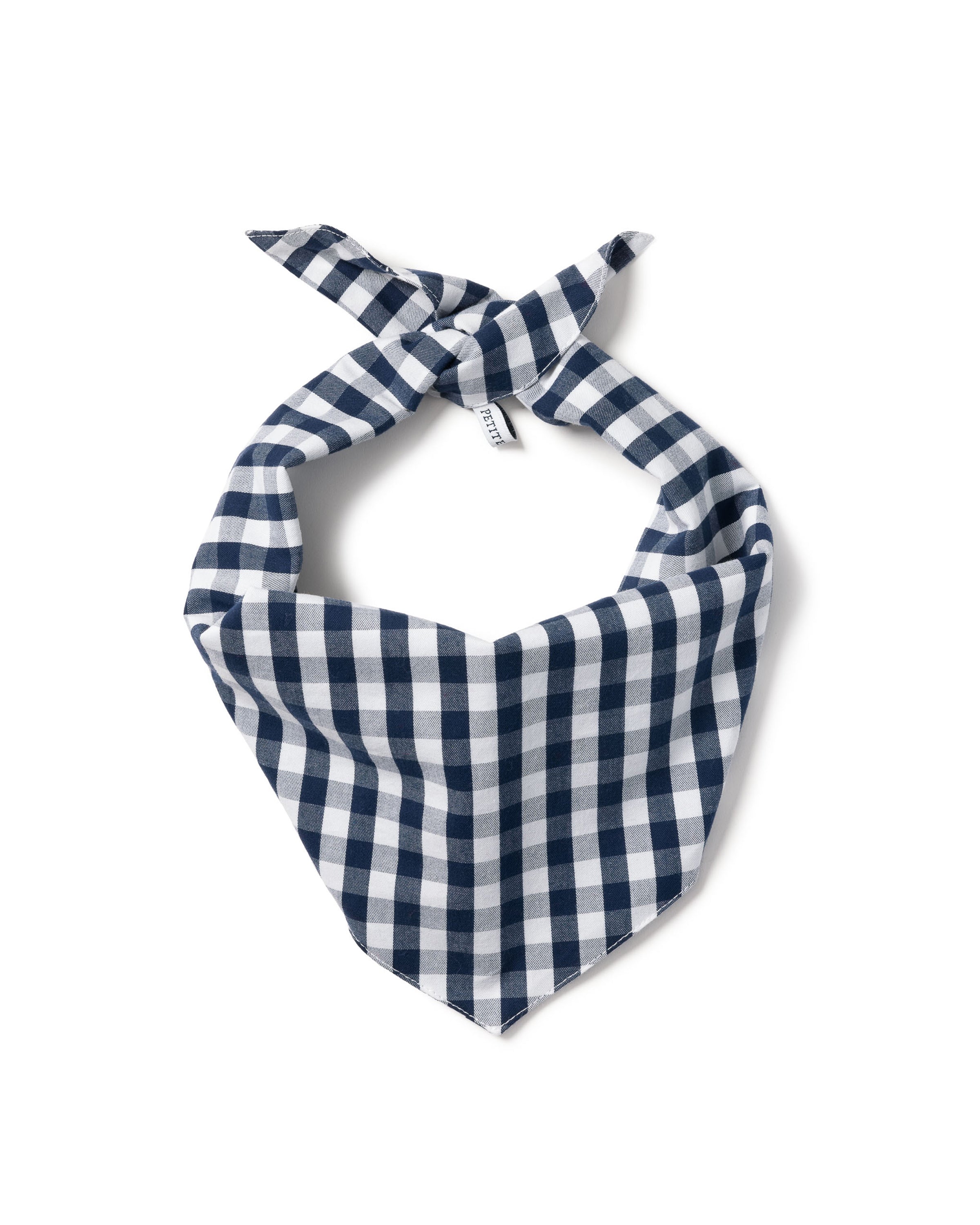 A fluffy white dog with pointy ears and a bushy tail sits on a wooden floor, wearing a Petite Plume Dog Bandana in Navy Gingham. It looks up with its tongue out, perfectly showcasing personality and style in its charming dog accessory.