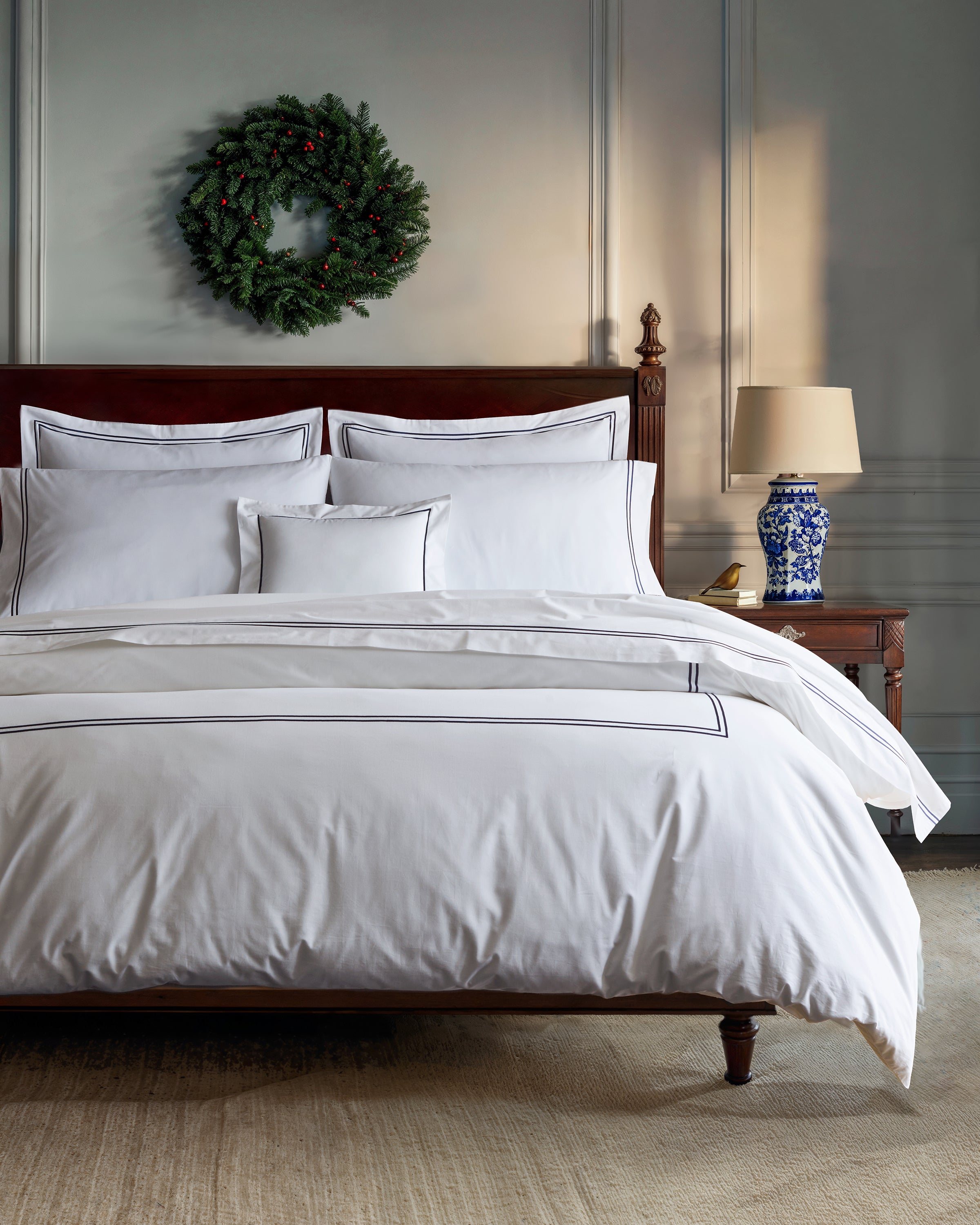 Elegant bedroom interior with a large bed featuring Petite Plumes Luxe Premium Percale Duvet Set in White with Navy Piping and a dark wood frame. A side table supports a lamp with a blue and white base, while a green wreath adds festivity to the light gray wall above the bed.