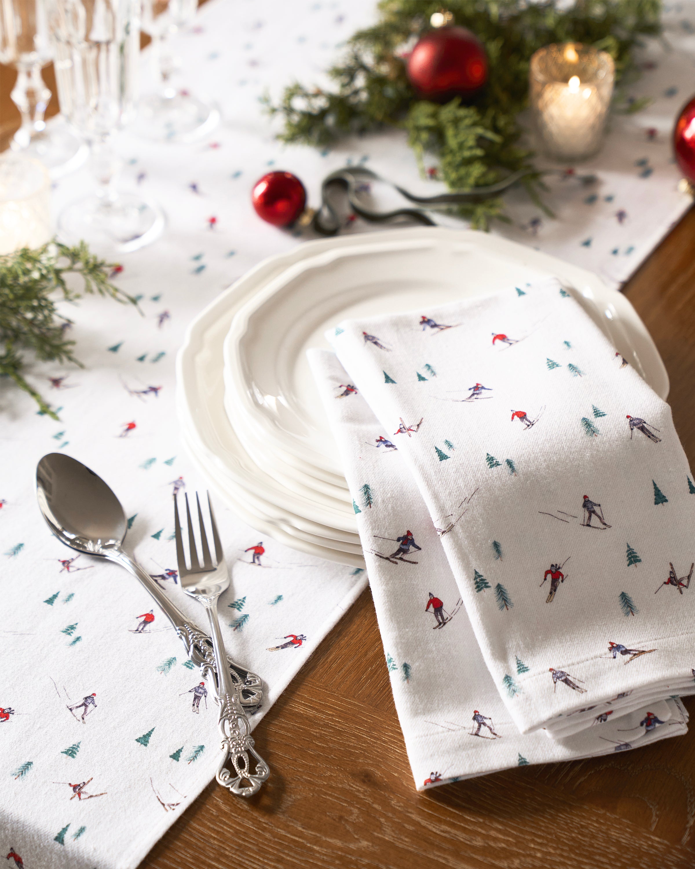 A table setting with white plates, a fork, and spoon on Petite Plumes Signature Twill Table Runner in Après Ski. Adorned with pine branches, red baubles, and lit candles on a wooden table, the décor crafts a festive tablescape exuding warmth and joy.